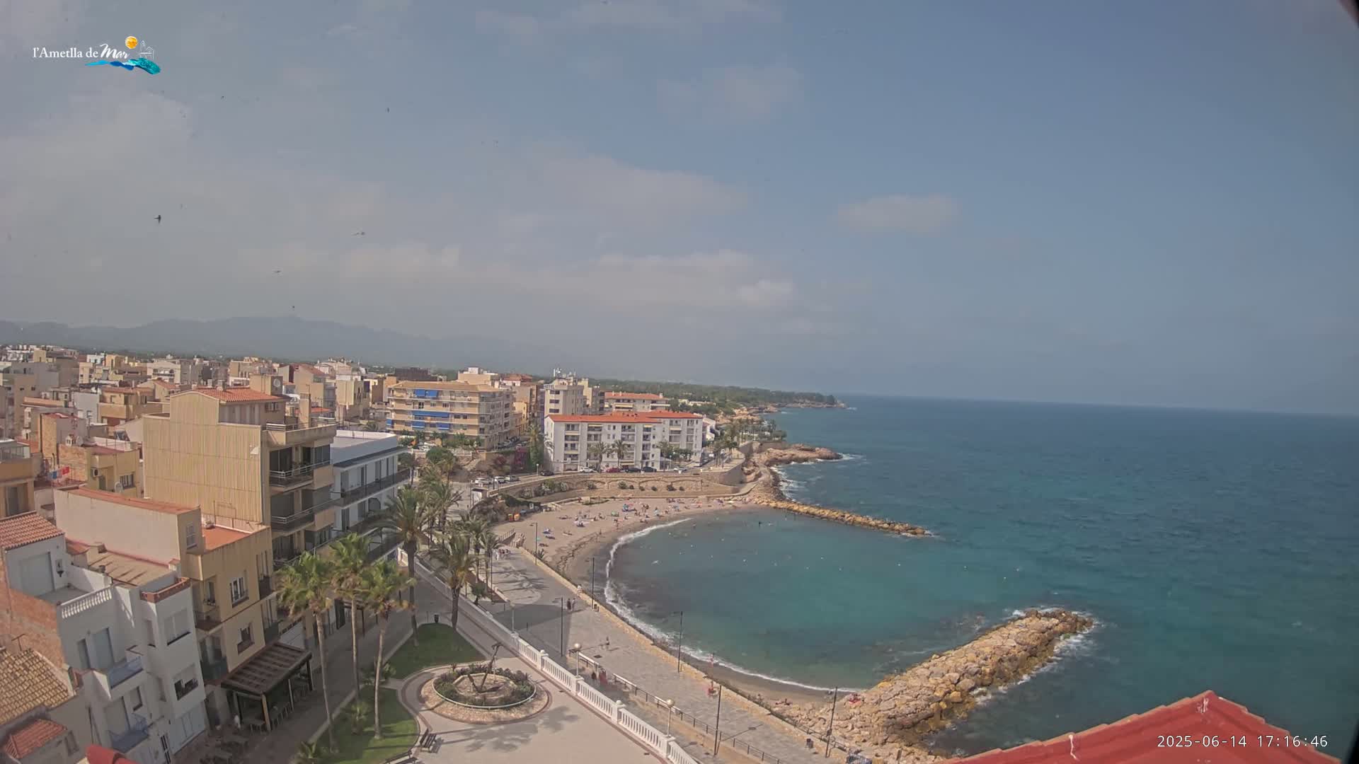 An elevated view shows a coastal town on a sunny day with a calm sea, a sandy beach with a few people, and buildings lining the shoreline.