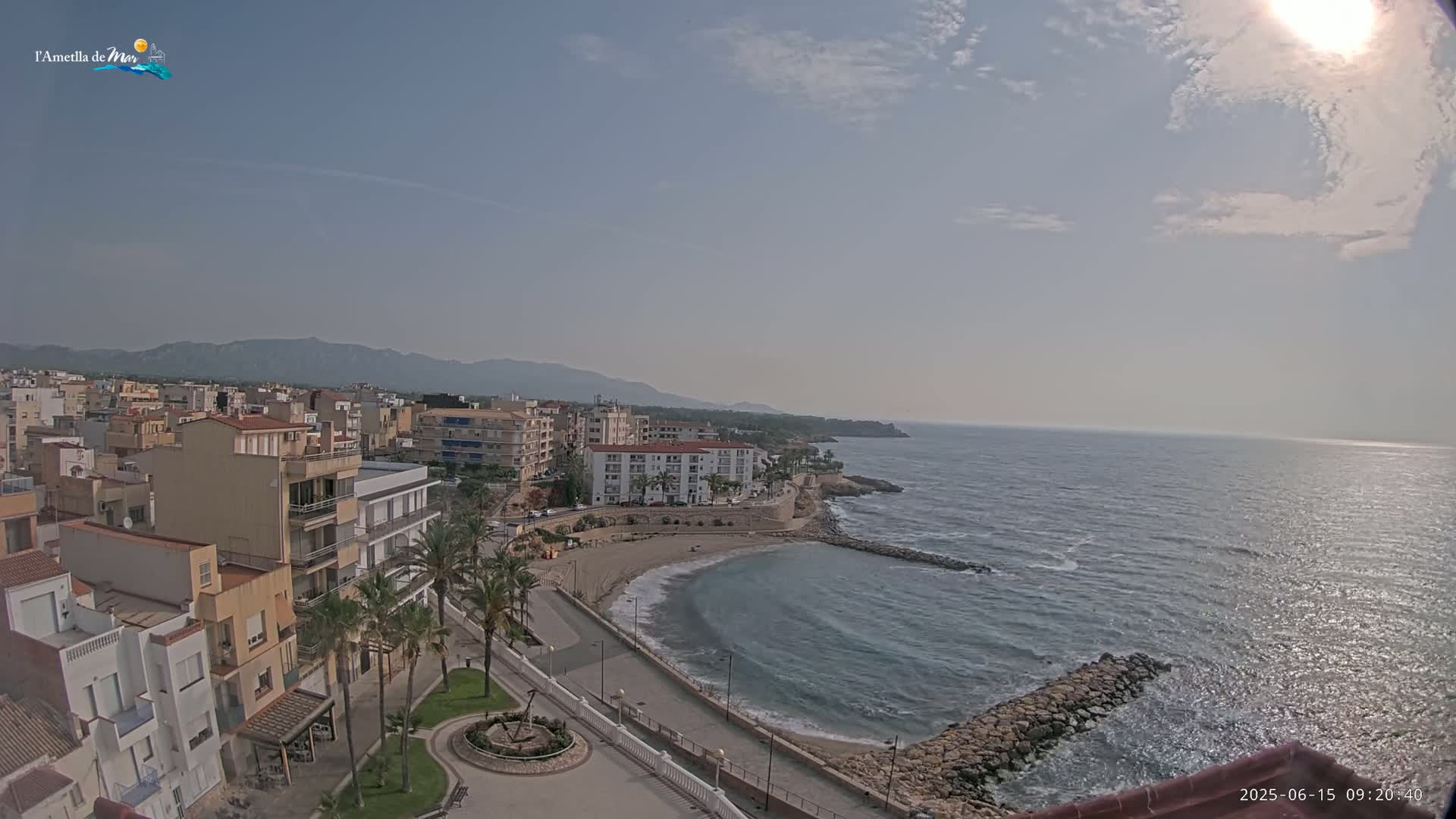 A sunny coastal town with a small sandy cove,  palm trees, and buildings stretches towards mountains in the distance under a mostly clear sky.