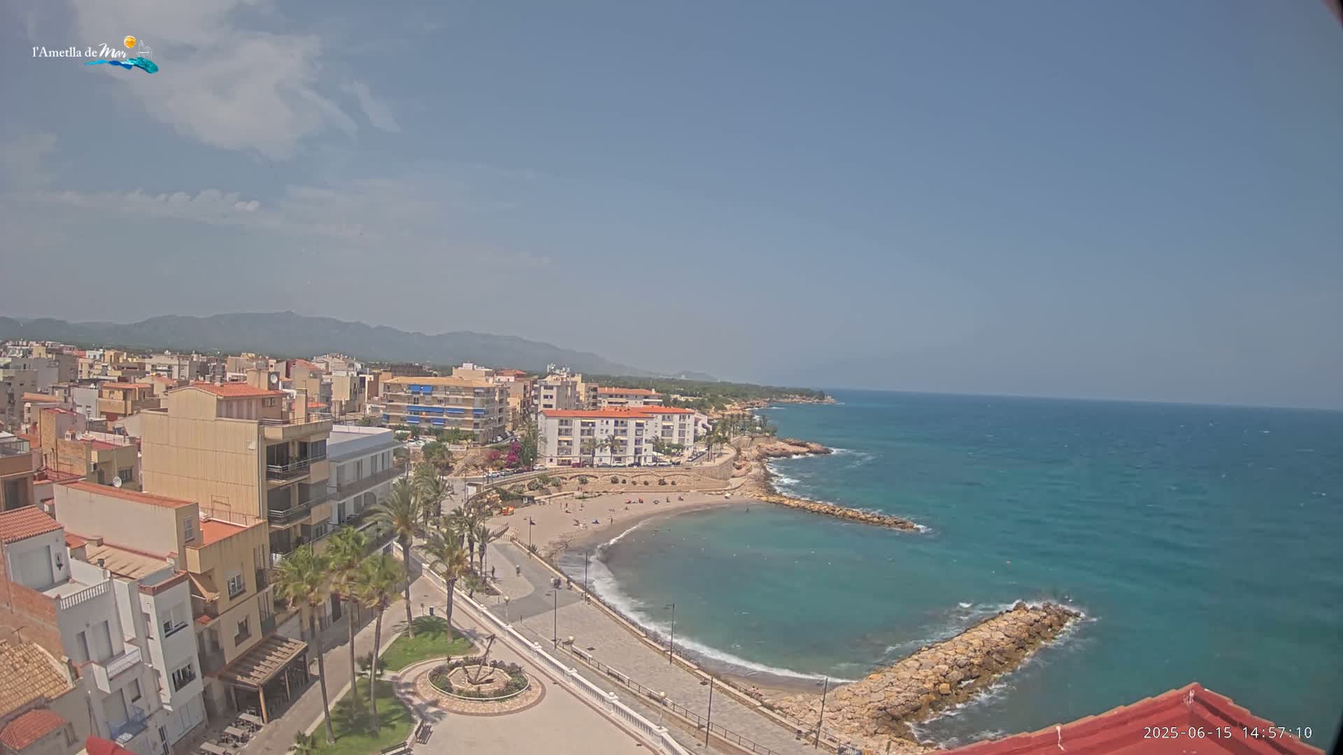 An aerial view shows a coastal town on a sunny day with a small, sandy beach curving into a calm, teal sea.