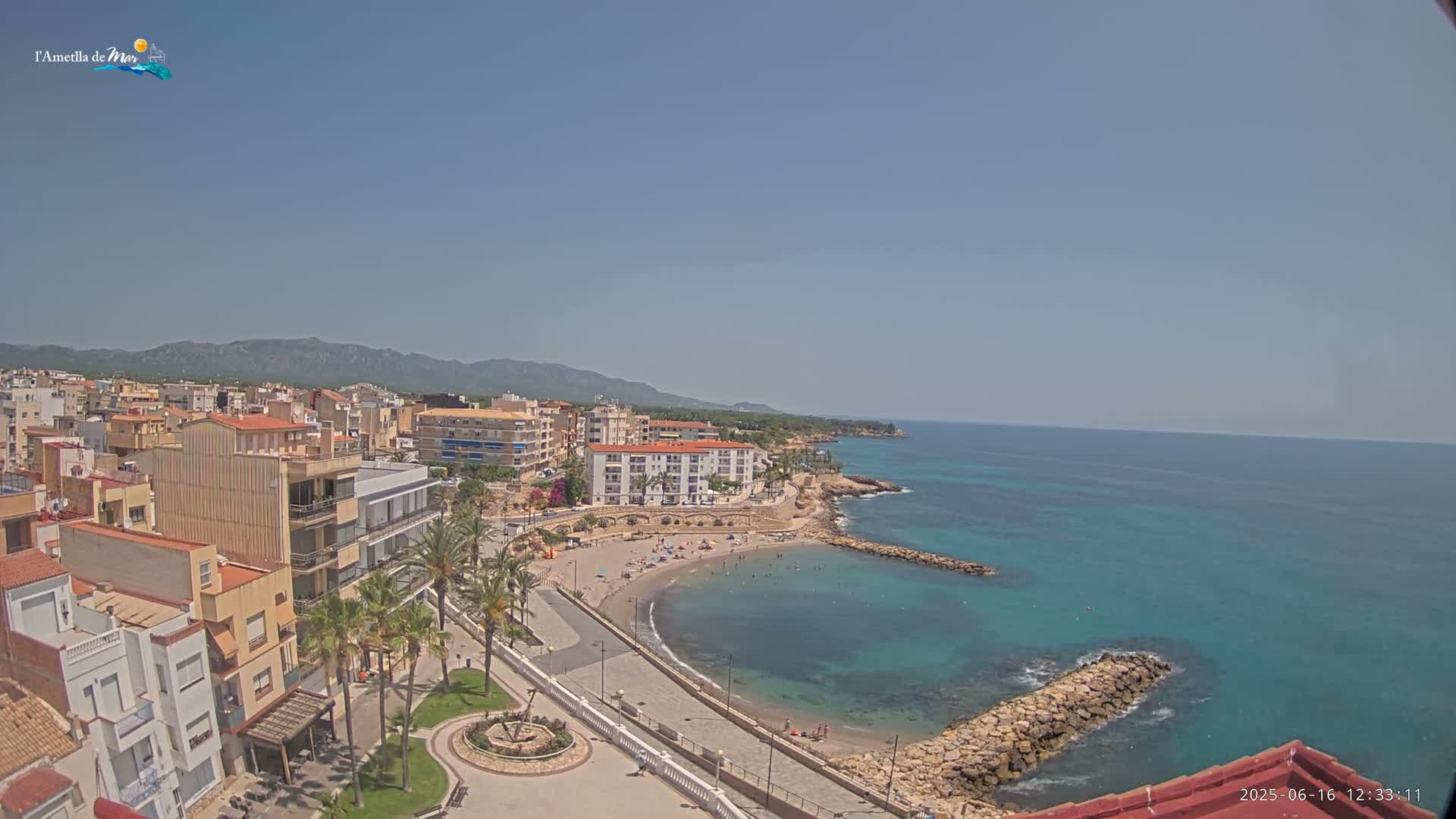 A coastal town with buildings, palm trees, a small sandy beach, and a calm, clear turquoise sea under a sunny sky.