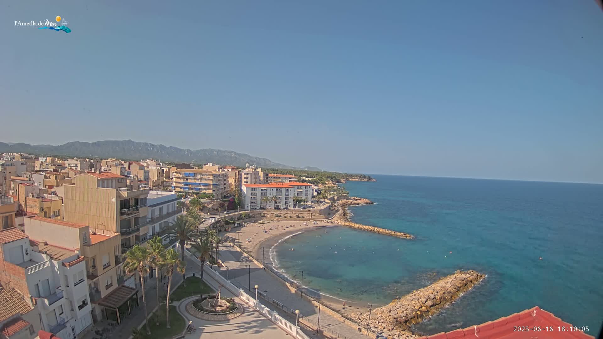 A coastal town with a sandy beach and calm turquoise water, backed by buildings and mountains under a clear blue sky.