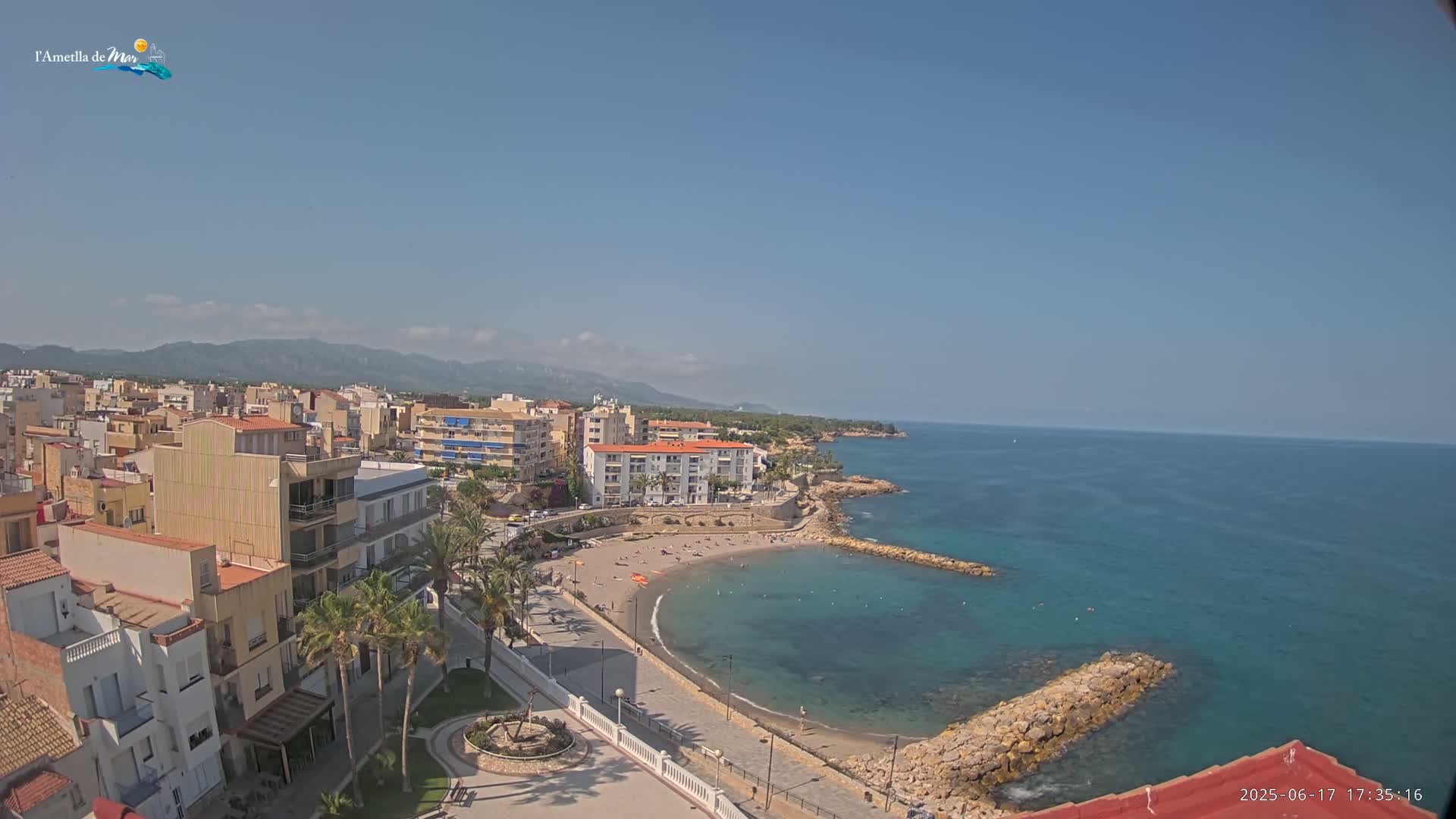 A coastal town with buildings, palm trees, and a small, crescent-shaped beach sits on a clear, sunny day next to a calm, teal-colored sea.