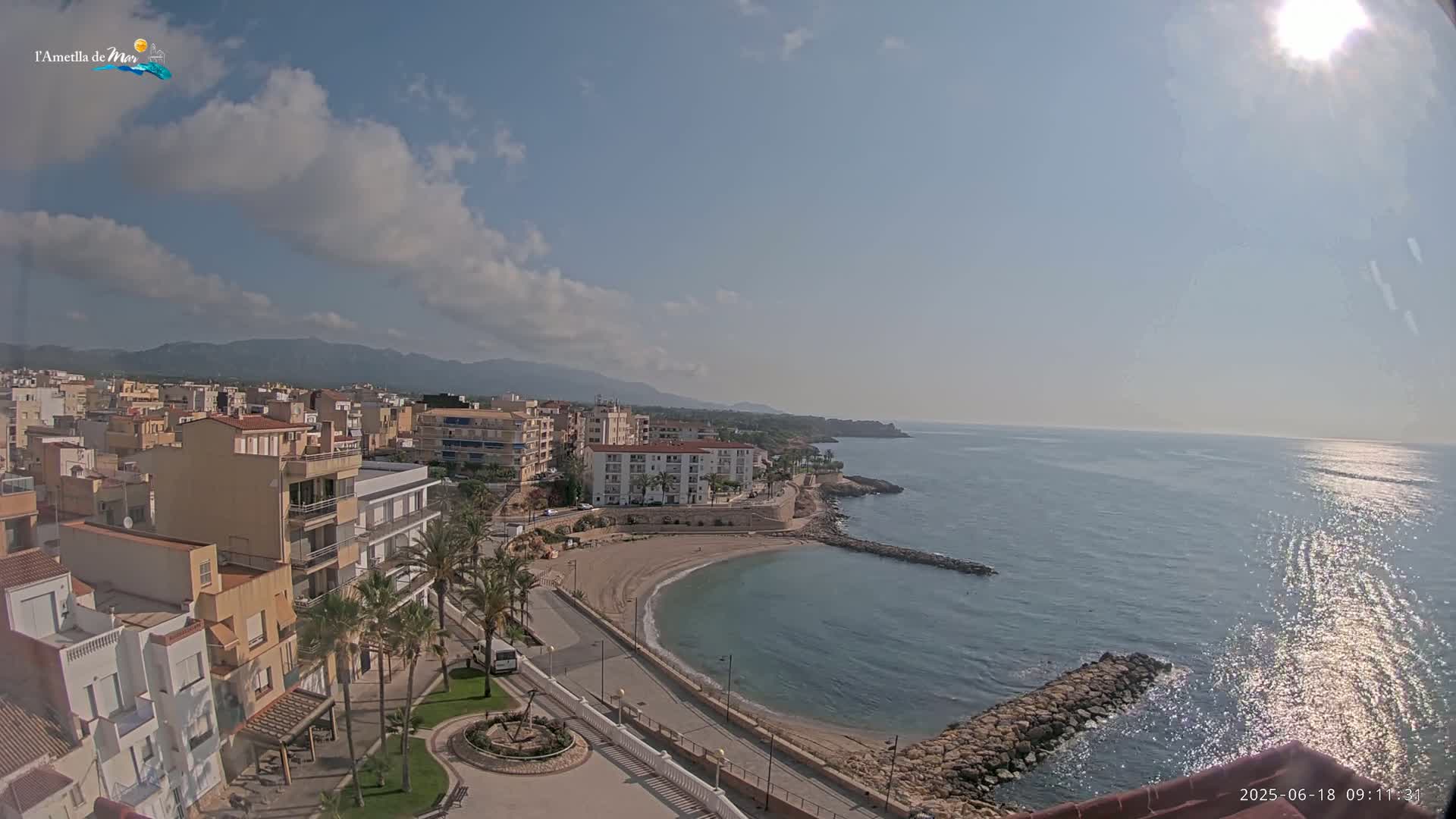 A partly sunny day reveals a coastal town with buildings, palm trees, a small sandy cove, and calm sea extending to distant mountains.