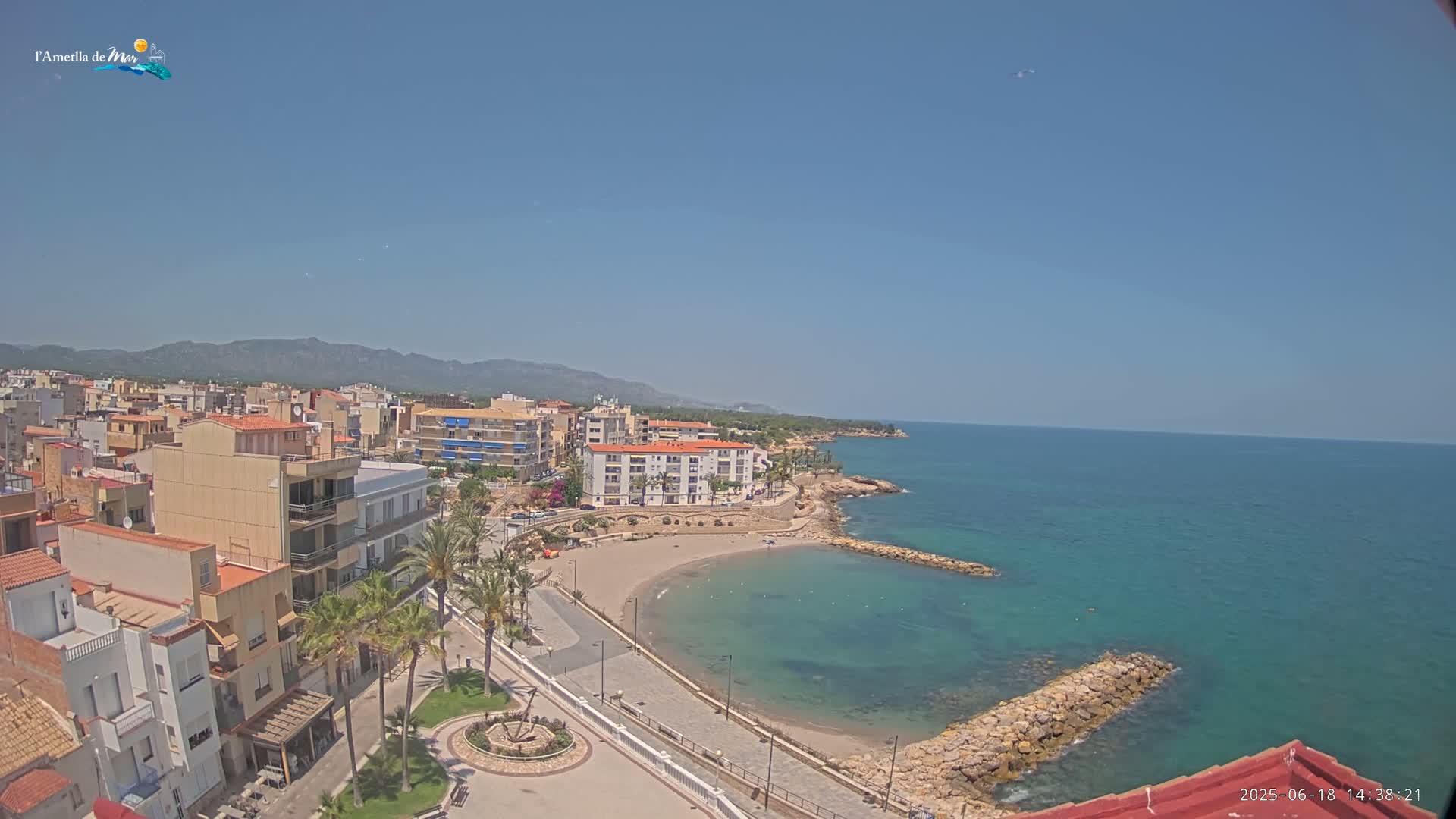 A coastal town with buildings, palm trees, a small sandy beach, and calm blue water under a clear blue sky.