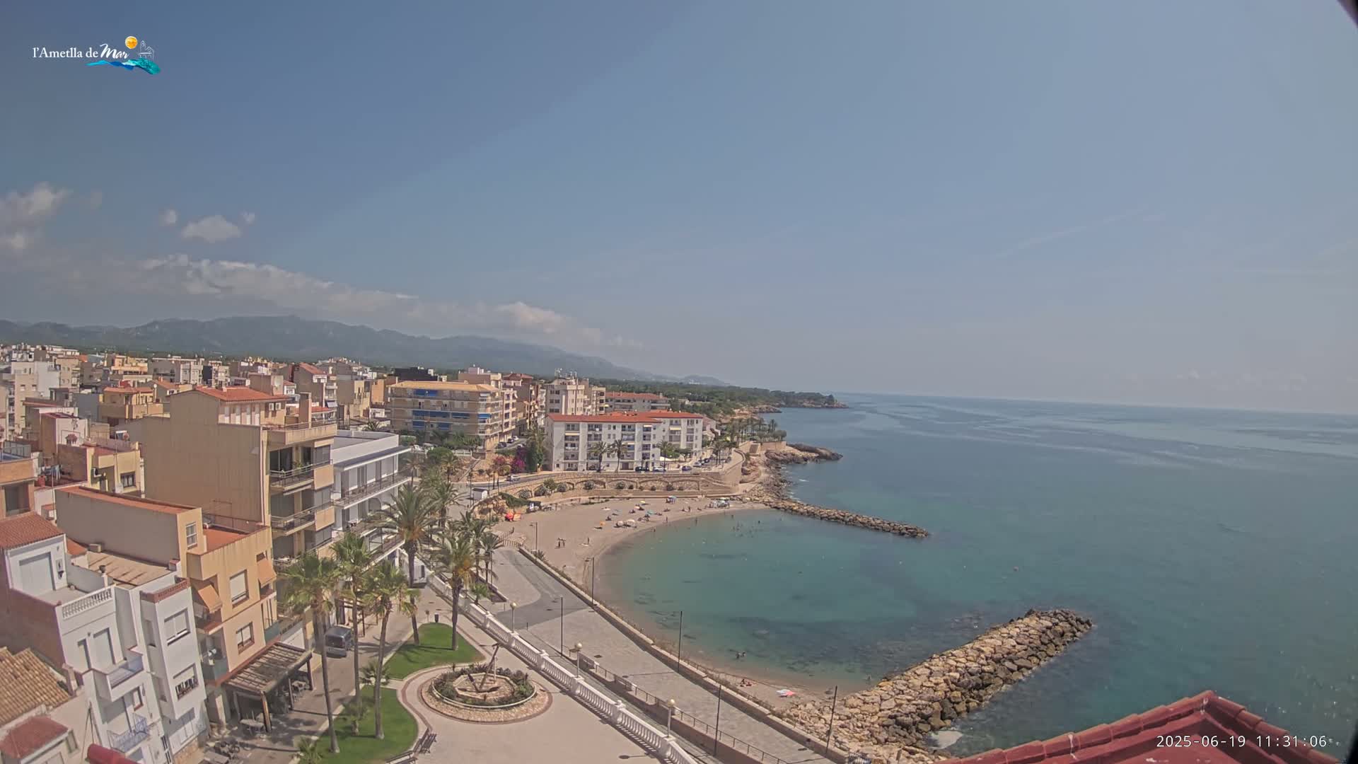 An aerial view shows a coastal town on a sunny day with a small, sandy beach curving into a calm, turquoise sea, backed by buildings and distant mountains.