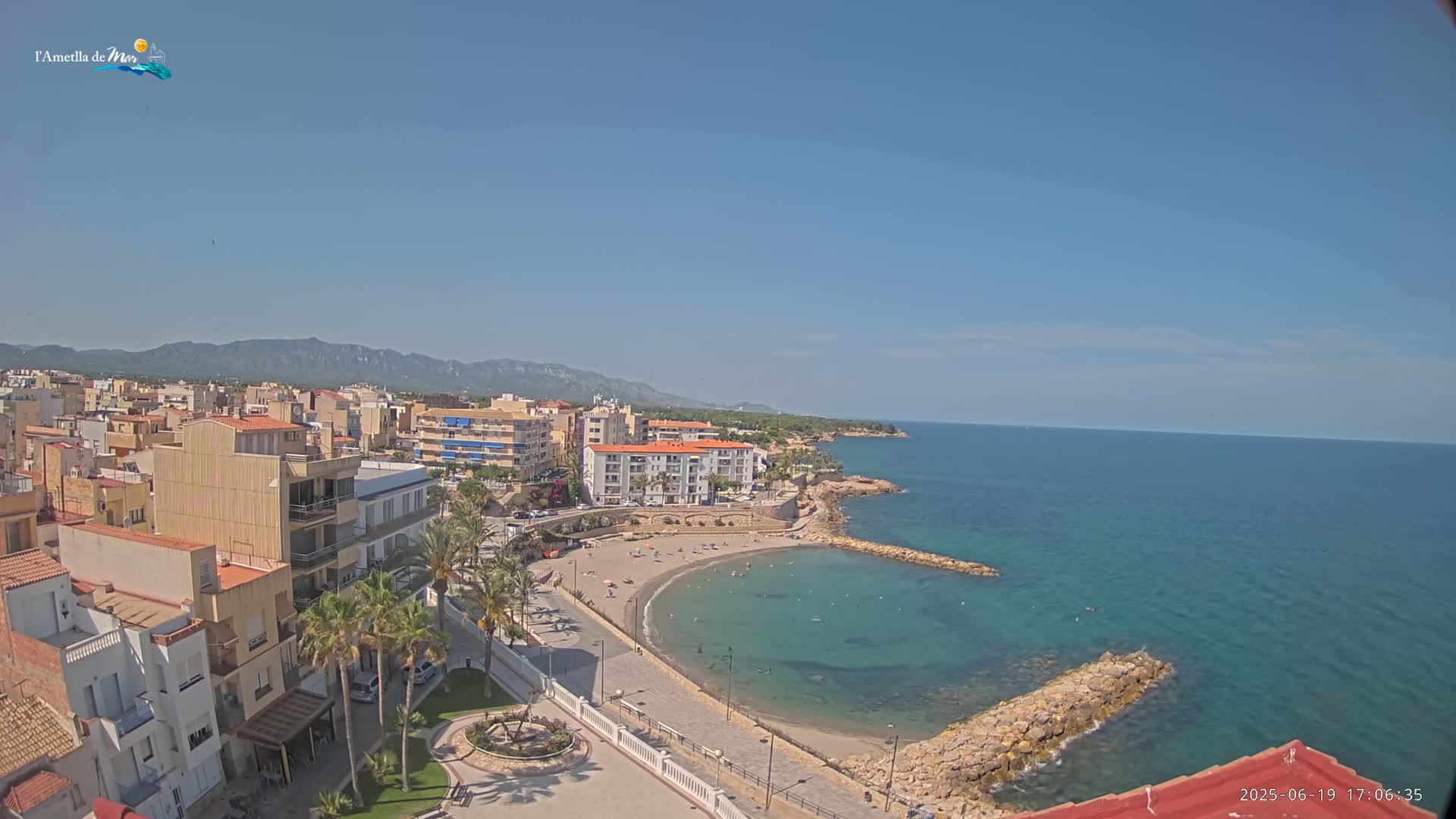 An aerial view shows a coastal town on a sunny day, with a small sandy beach curving into a calm, clear turquoise bay, and mountains visible in the distance.