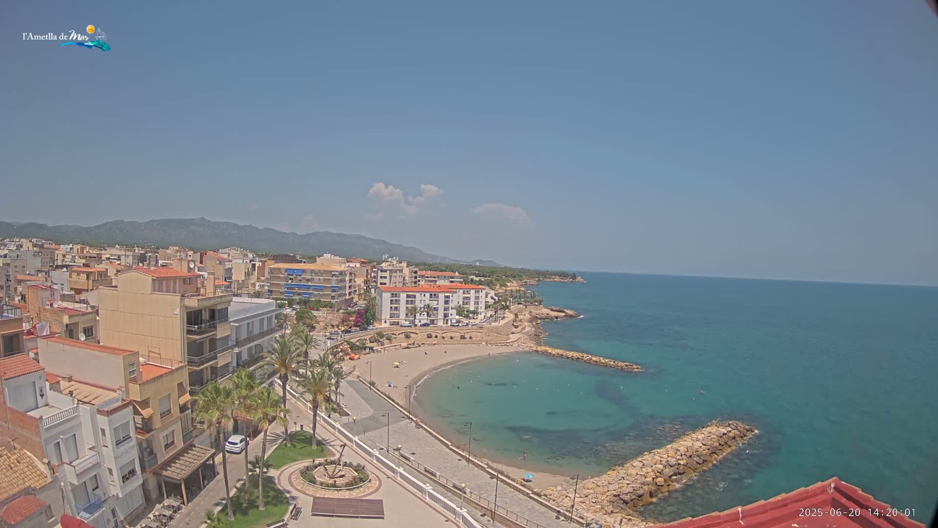 A coastal town with low-rise buildings and a small, sandy beach nestled into a calm, teal sea under a mostly clear sky.