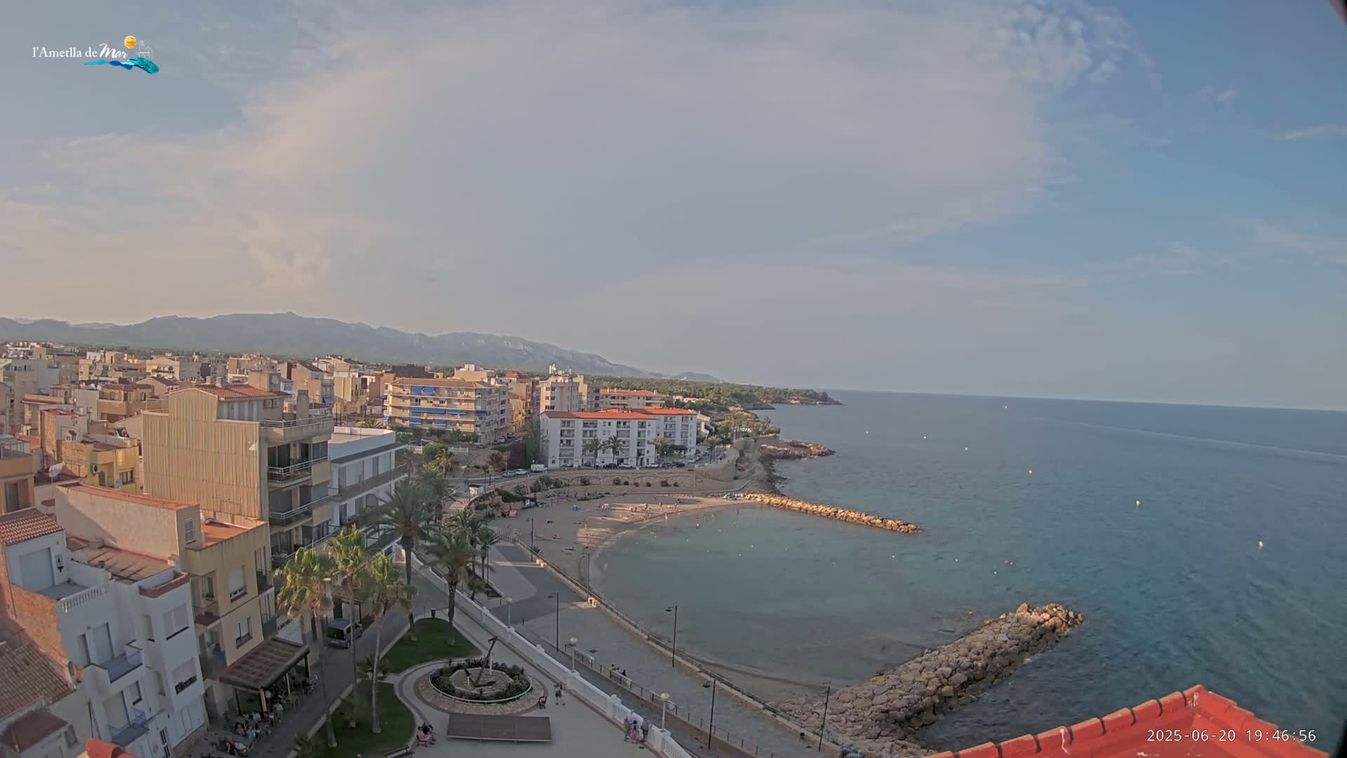 An aerial view shows a partly sunny coastal town with a small beach and calm ocean, backed by low mountains.