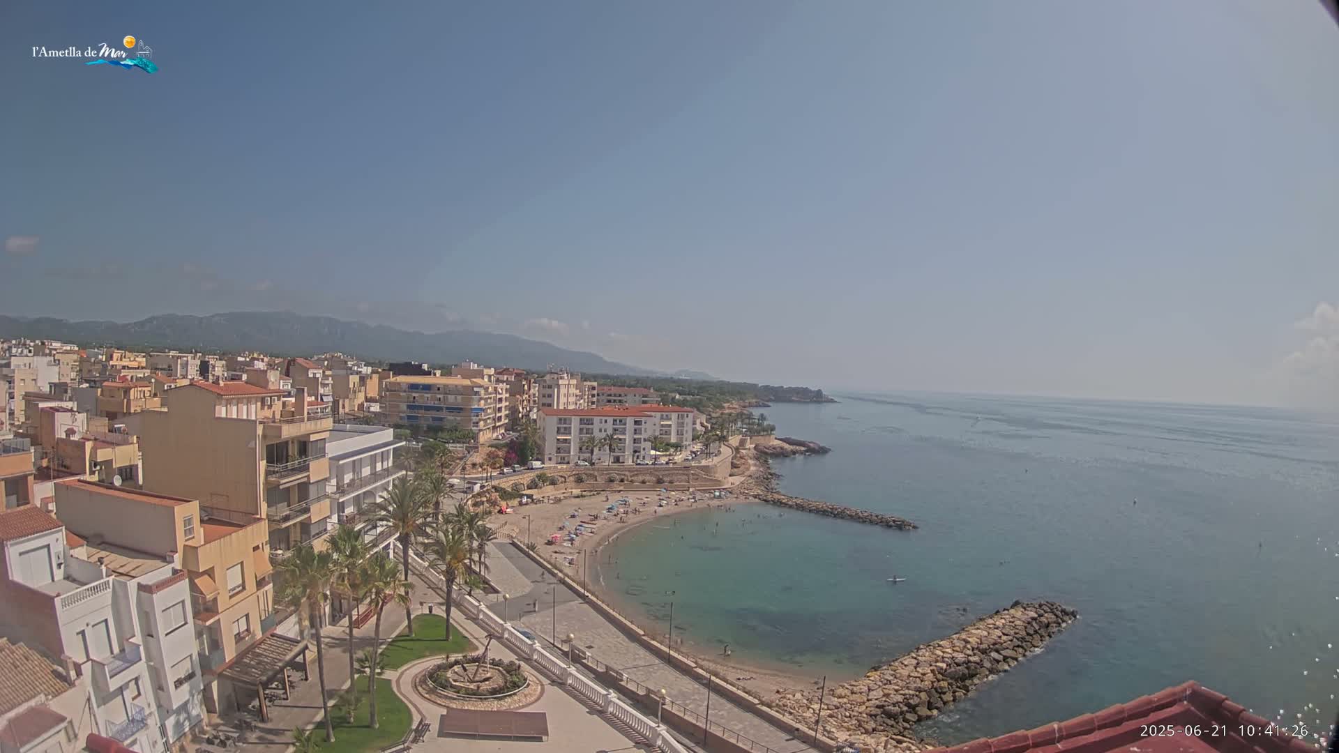 A coastal town with a small, sandy beach and calm, clear water is seen on a sunny day, with mountains visible in the distance.