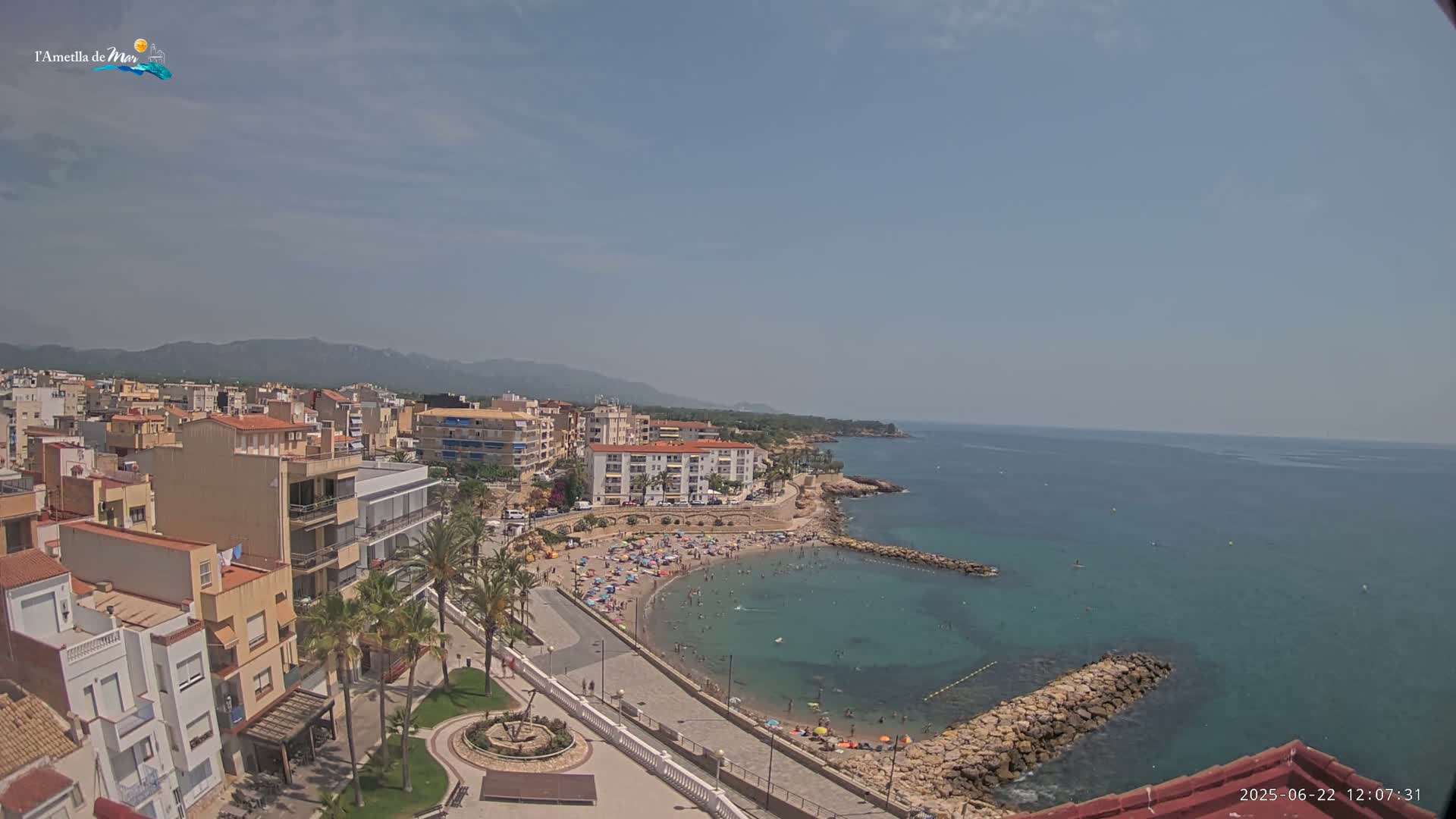 A sunny day reveals a coastal town with a crowded beach, calm blue water, and a rocky shoreline, backed by buildings and mountains.