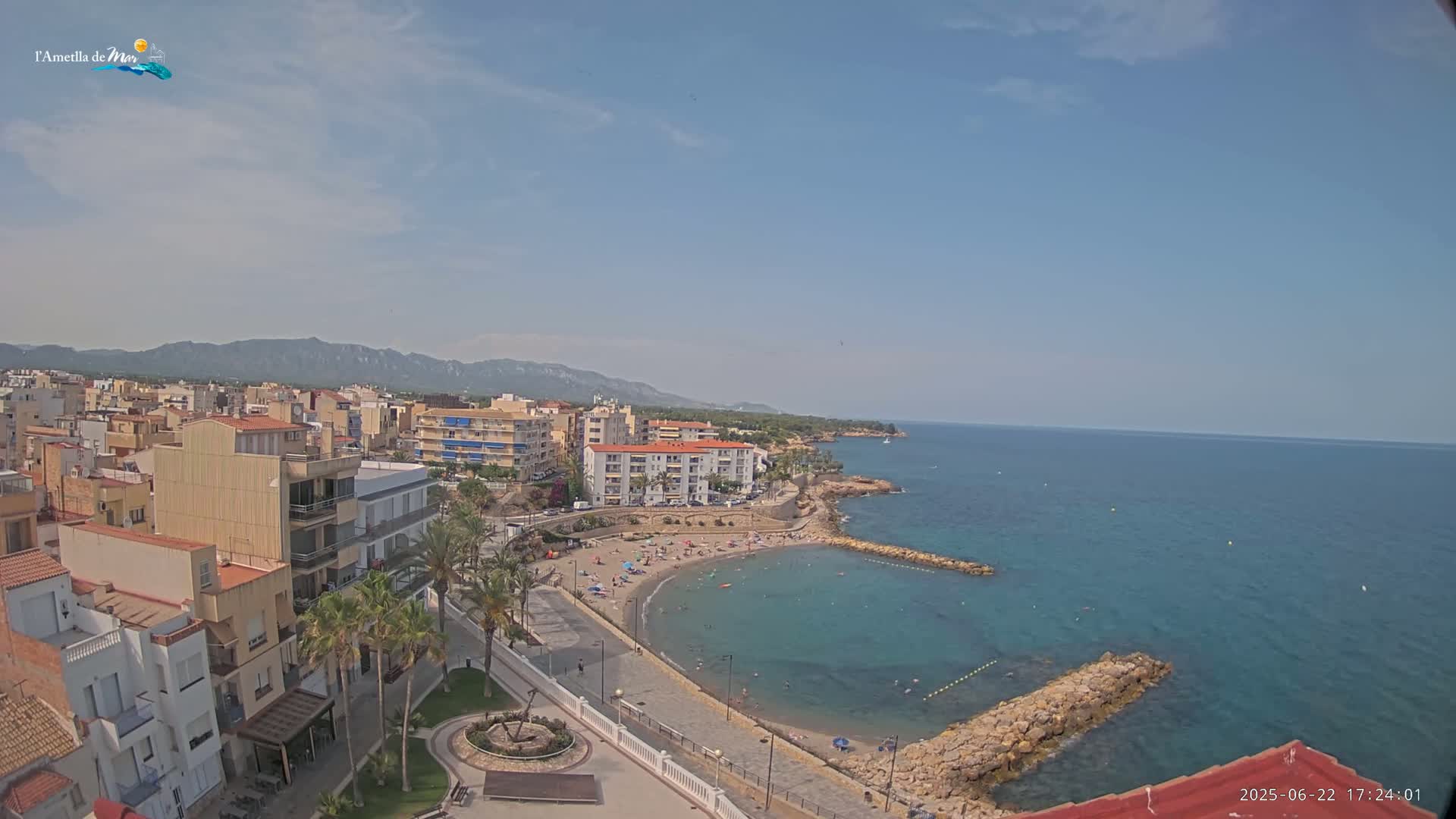 An elevated view shows a coastal town on a sunny day with a calm blue sea, a sandy beach with people, and buildings stretching into the background towards mountains.