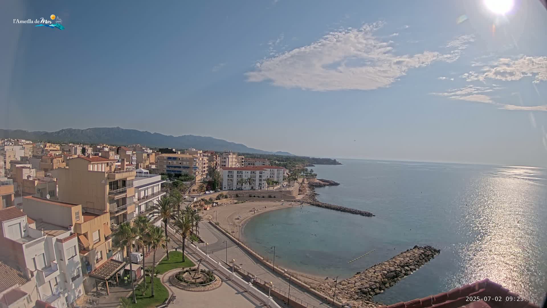 A sunny day reveals a coastal town with buildings, a small crescent-shaped beach, calm blue water, and mountains in the distance.