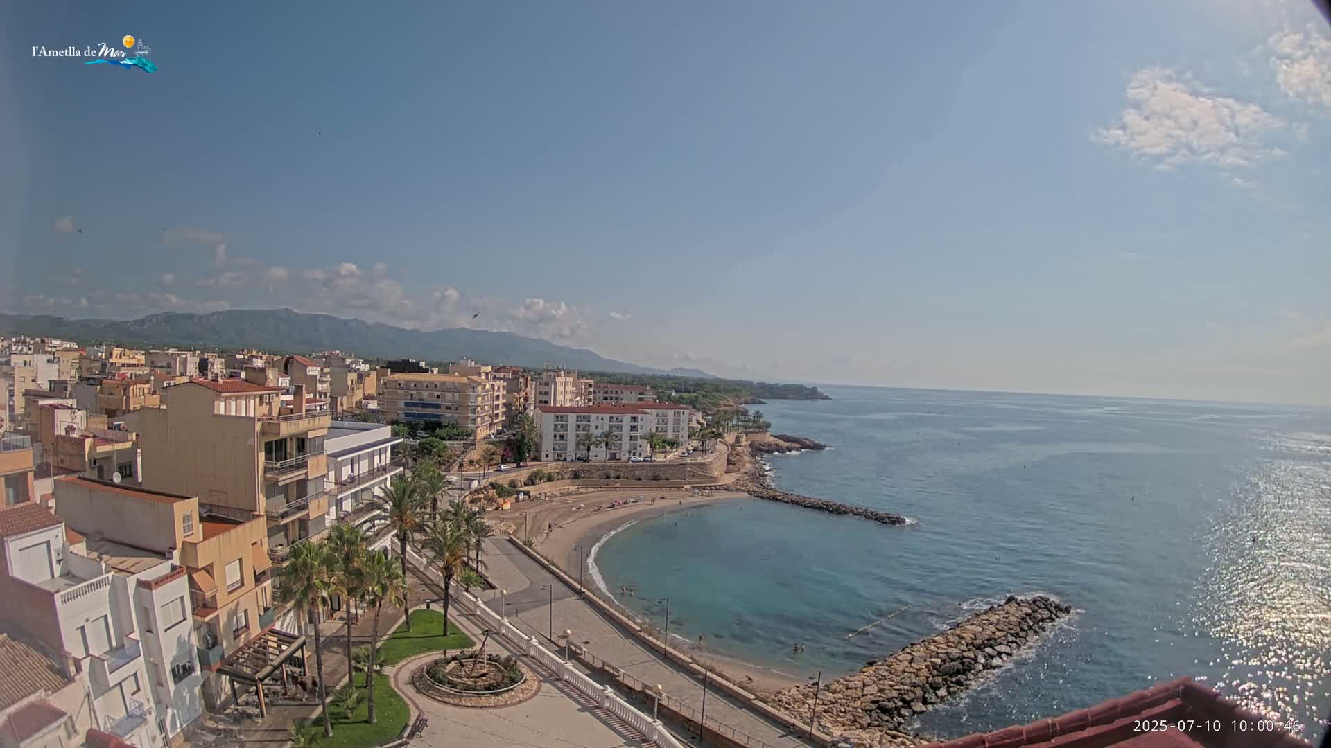 A coastal town with buildings, palm trees, a small beach, and calm blue water under a mostly sunny sky.