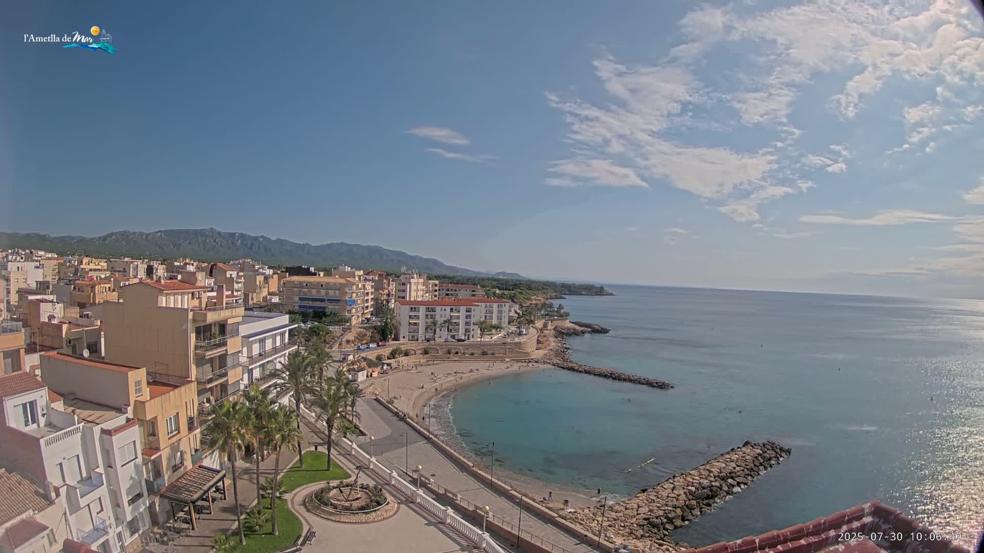 A partly cloudy sunny day overlooks a coastal town with a small beach and calm, clear turquoise water, backed by mountains.