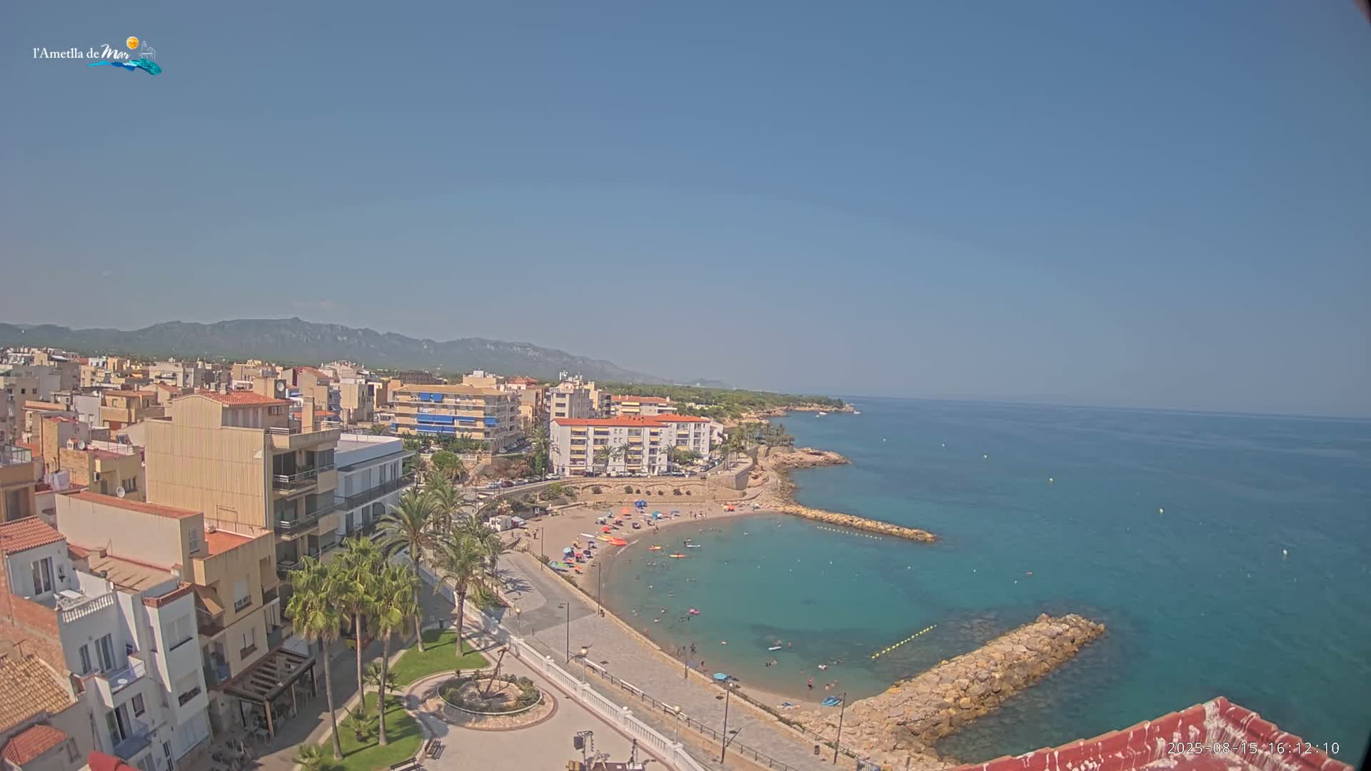 A partly cloudy sunny day overlooks a coastal town with a small beach and calm, clear turquoise water, backed by mountains.