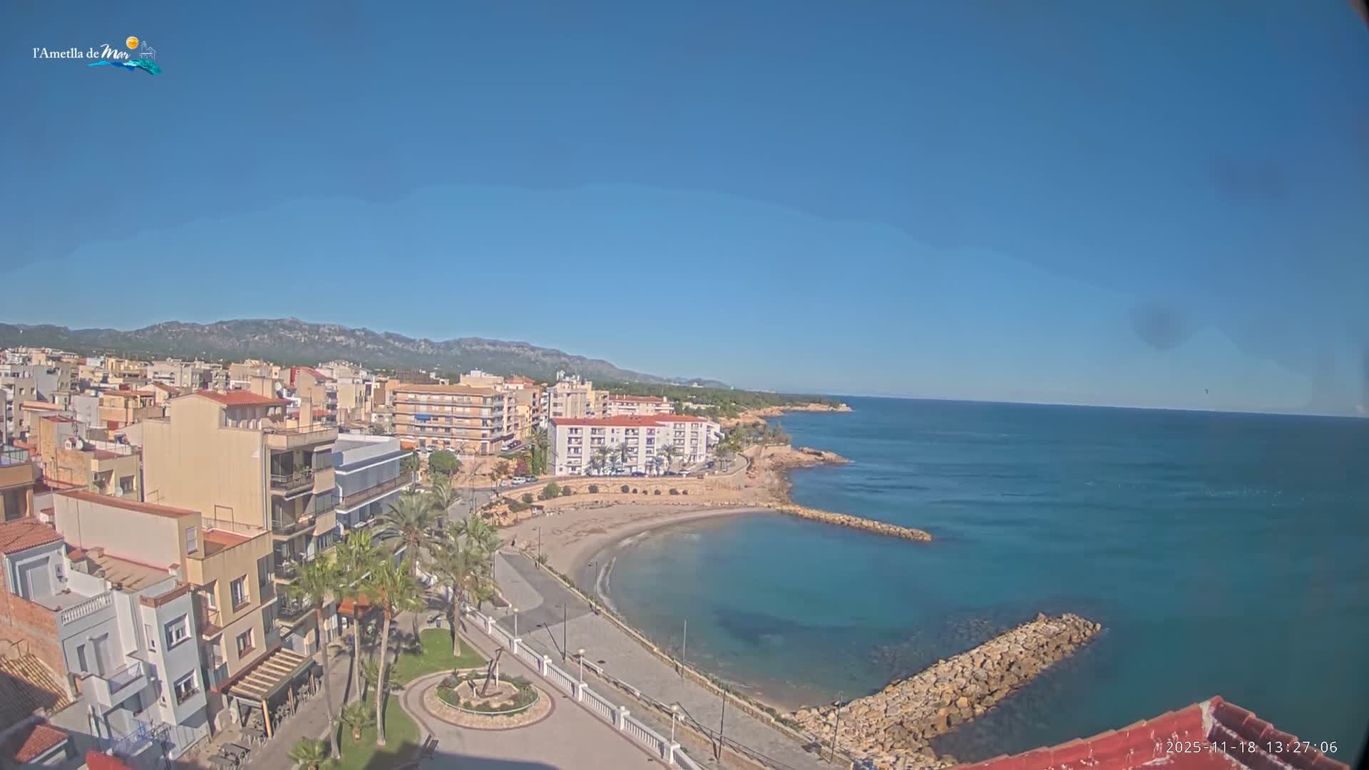 A partly cloudy sunny day overlooks a coastal town with a small beach and calm, clear turquoise water, backed by mountains.
