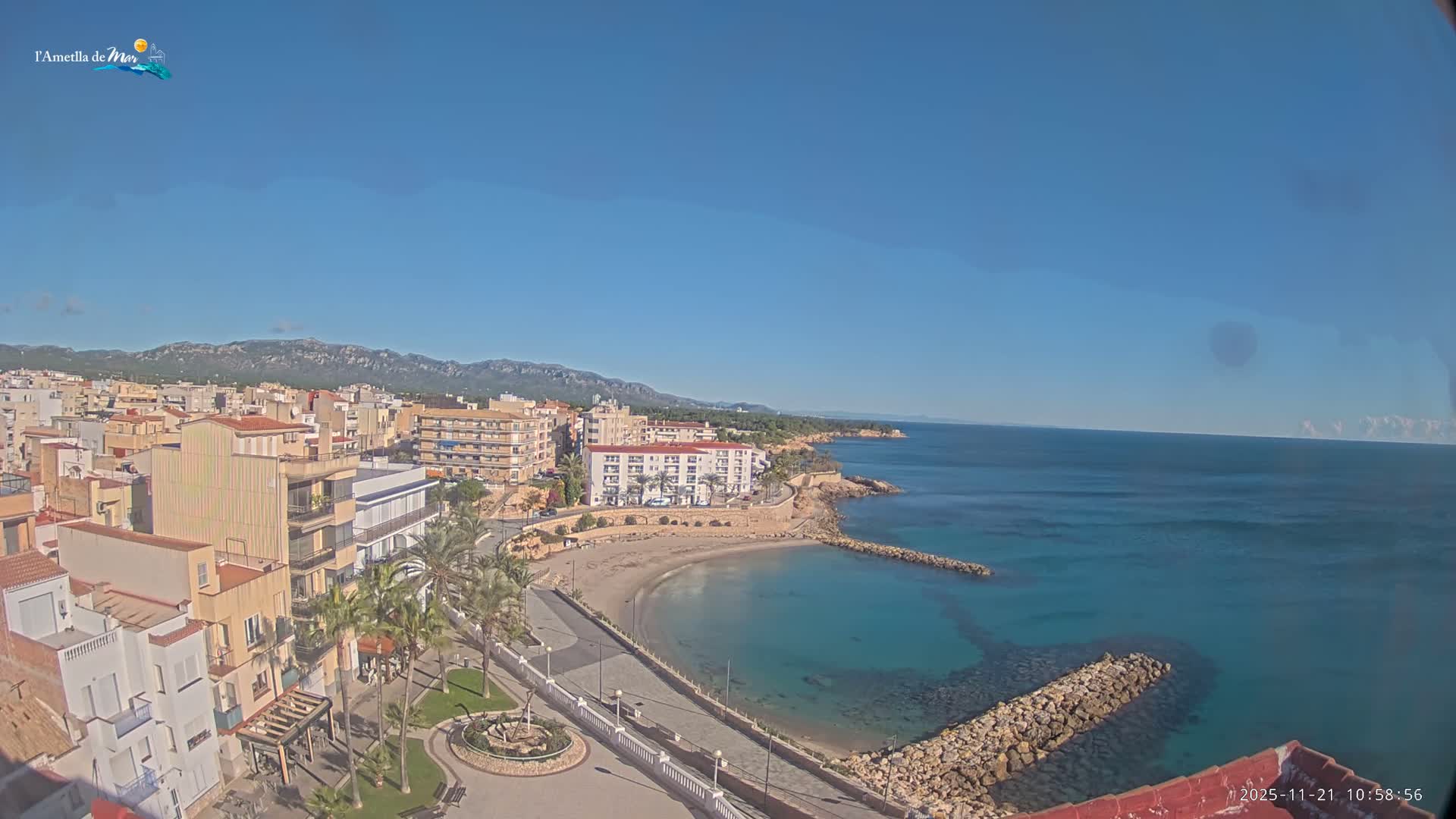 Under a clear blue sky, a coastal town features buildings lining a promenade with palm trees, a curved sandy beach, and calm turquoise waters with rock jetties, backed by distant mountains.