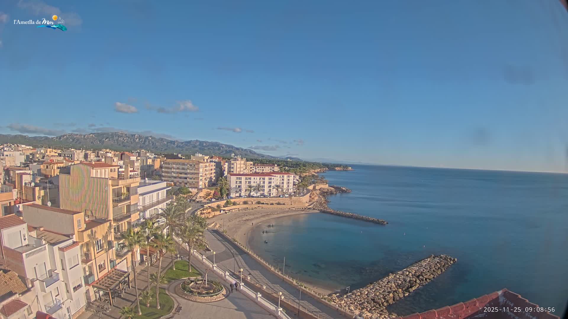 An elevated view captures a sunny coastal town with numerous buildings, palm-lined streets, a curved sandy beach with stone breakwaters, and a calm blue sea, all under a clear sky with a few scattered clouds, backed by distant mountains.