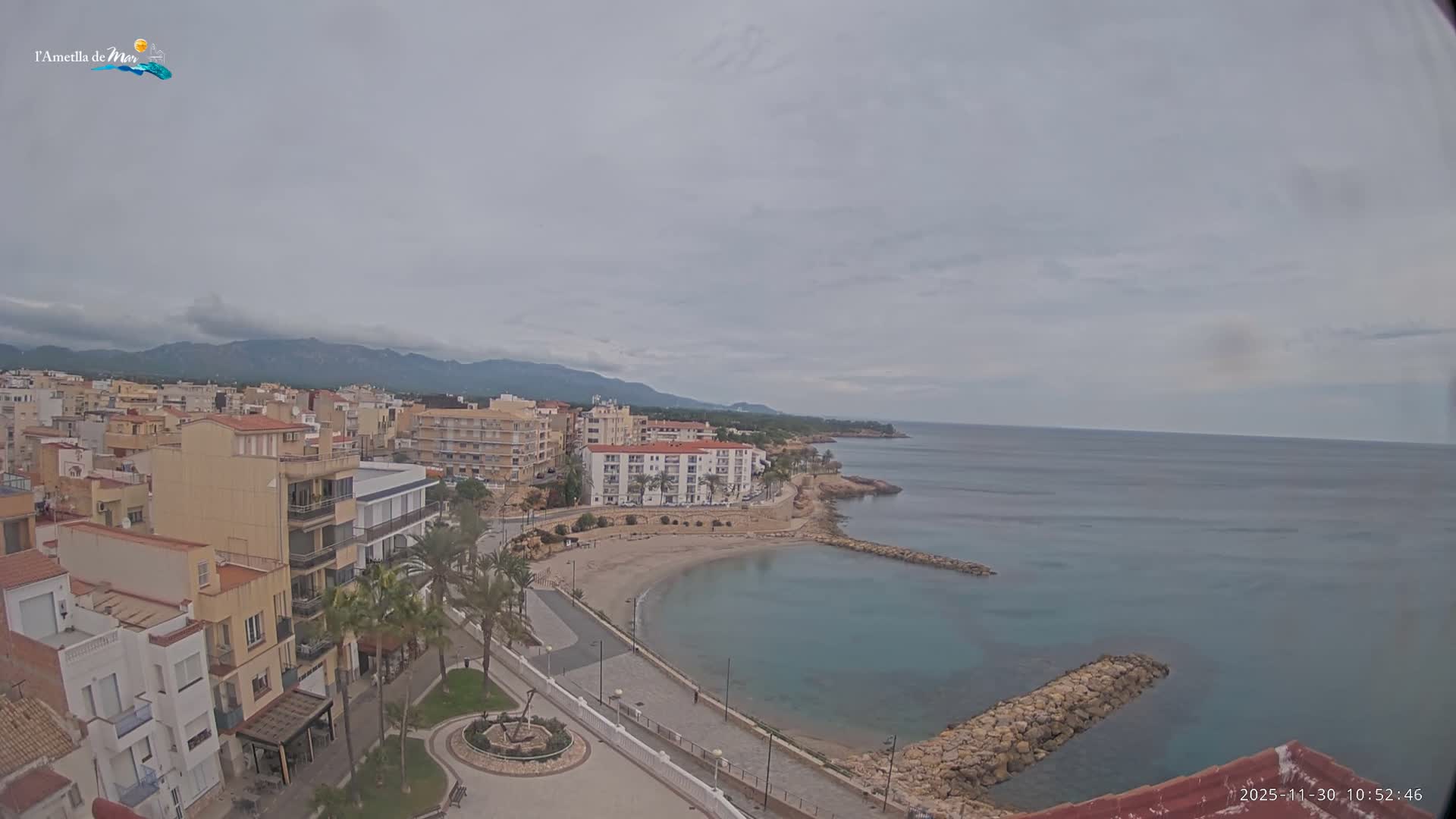An elevated view reveals a coastal town featuring buildings lining a promenade, a sandy beach with two stone breakwaters, and calm turquoise waters stretching to the horizon, all beneath an overcast sky with distant mountains visible.