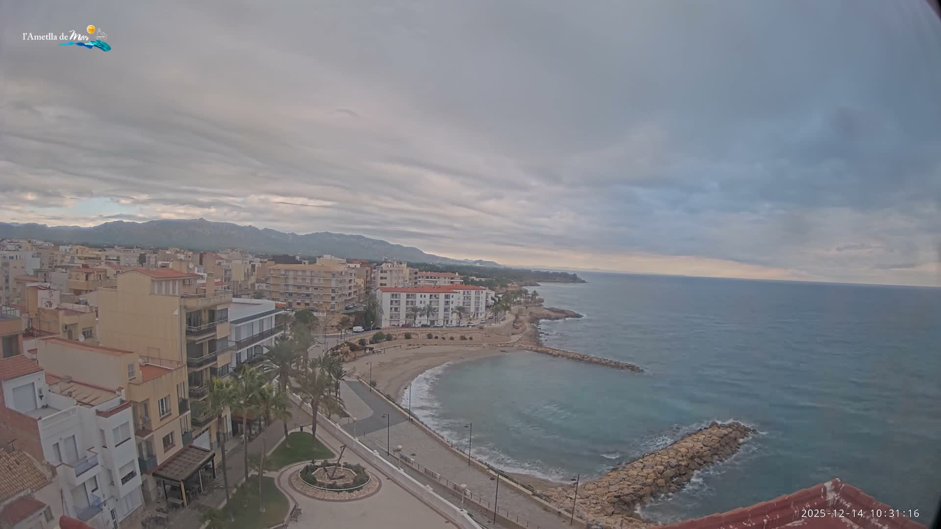 An elevated view reveals a coastal town featuring buildings lining a promenade, a sandy beach with two stone breakwaters, and calm turquoise waters stretching to the horizon, all beneath an overcast sky with distant mountains visible.