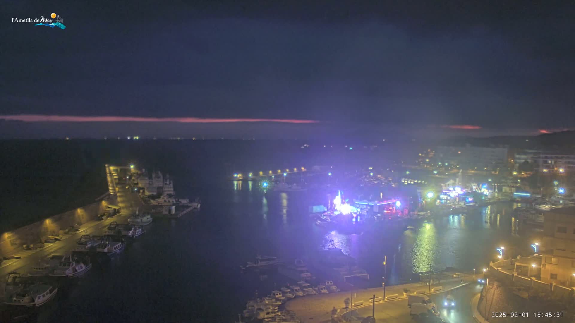 A nighttime aerial view of a harbor filled with boats, with a brightly lit waterfront area and a hazy, pinkish sky on the horizon.