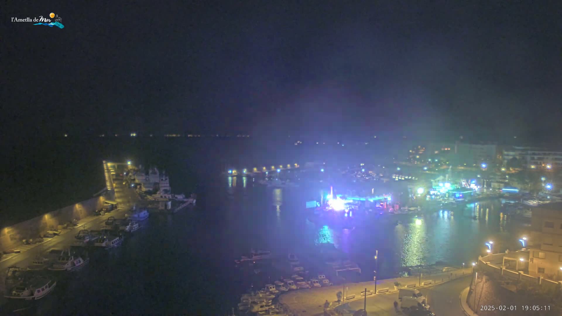 A nighttime aerial view of a harbor with boats docked along a lit waterfront, a brightly lit event or show taking place on the water, and a hazy atmosphere.