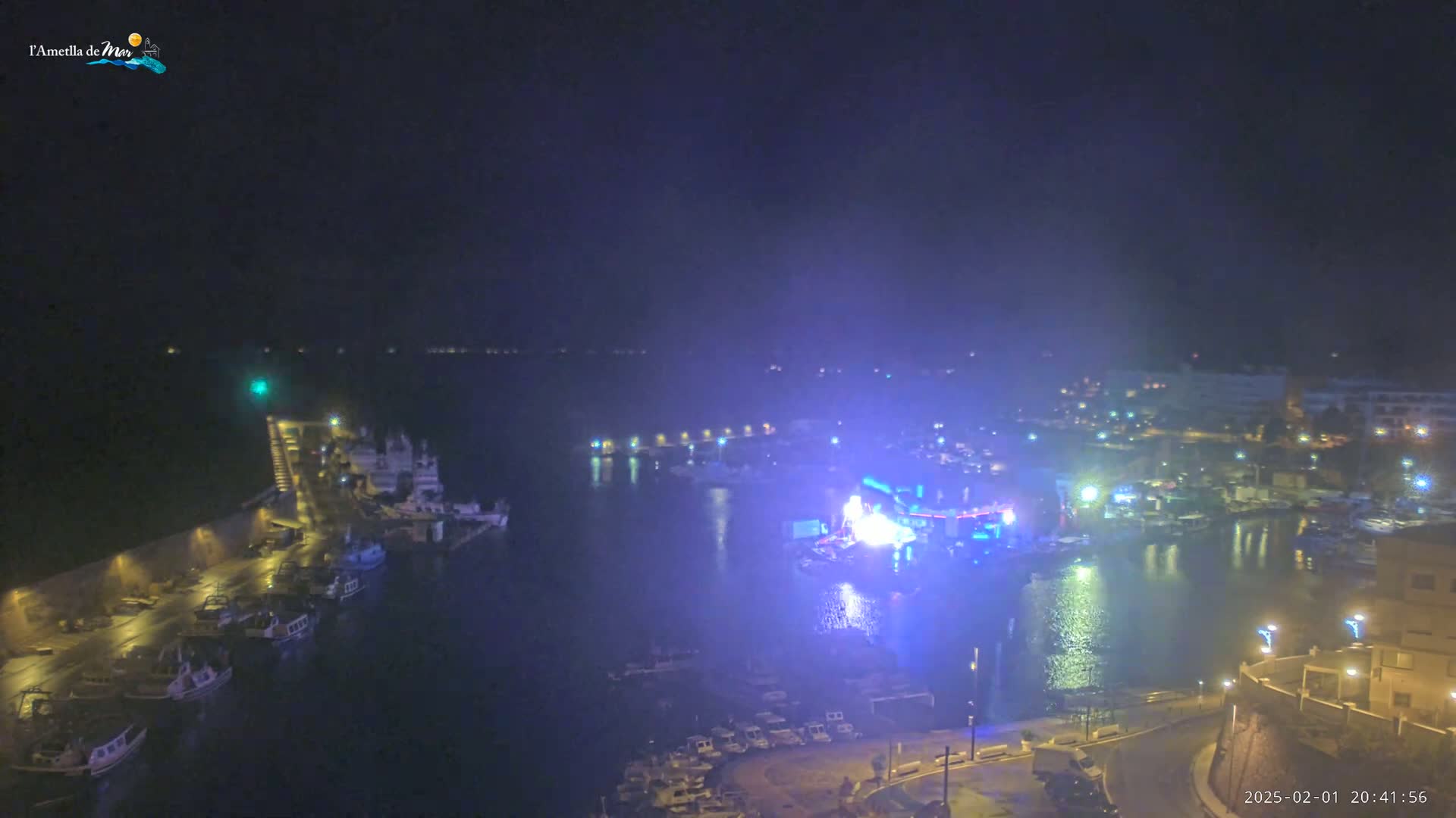 A nighttime aerial view of a harbor with boats and a brightly lit stage or performance area, under hazy or foggy conditions.
