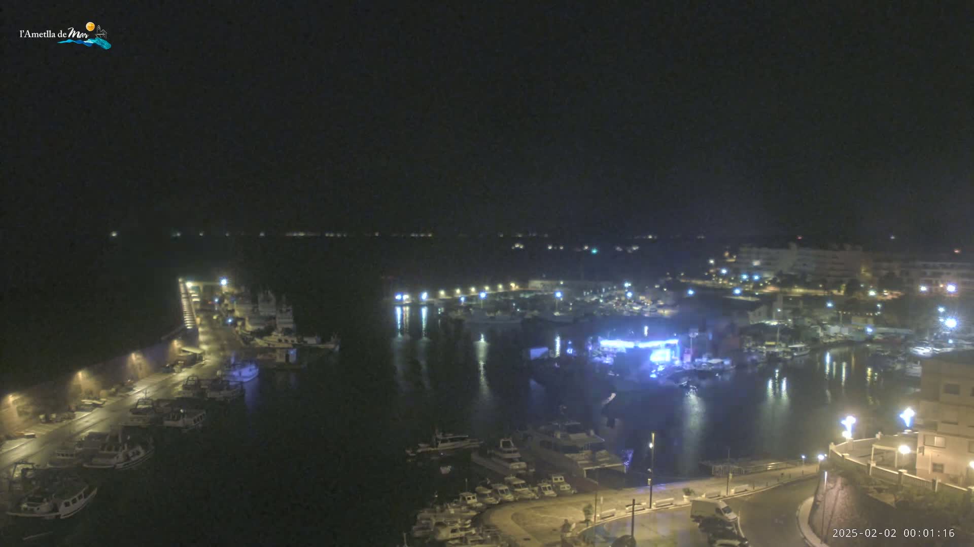 A nighttime view of a harbor filled with boats, illuminated by lights along the waterfront under a dark sky.