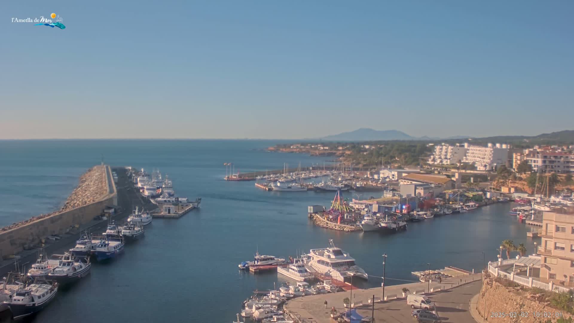 A harbor filled with numerous boats and ships is seen on a sunny day, with calm water and a distant mountain range visible on the horizon.