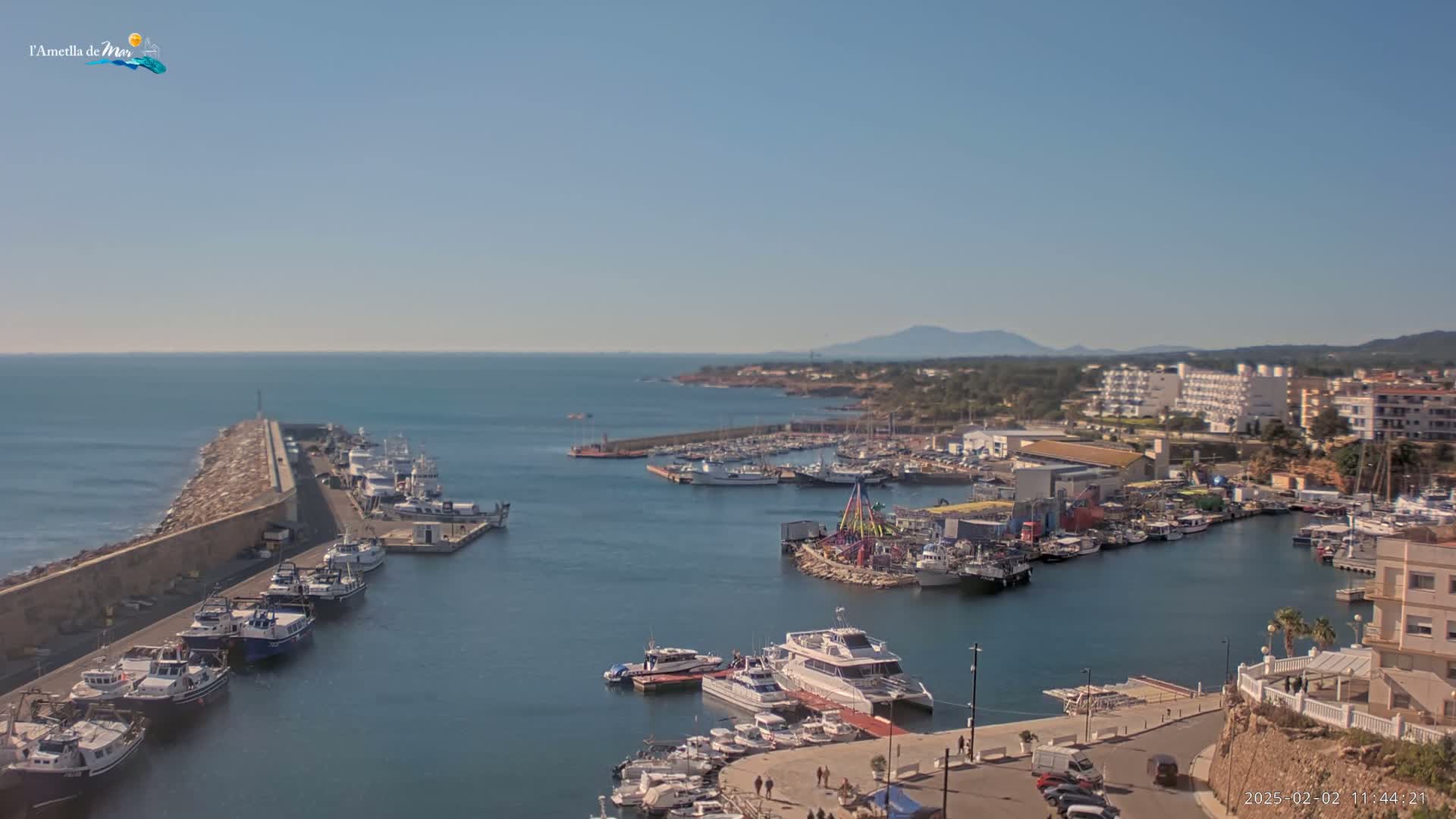 A harbor filled with numerous boats and ships is shown on a sunny day, with a calm sea extending to a distant coastline and mountains.