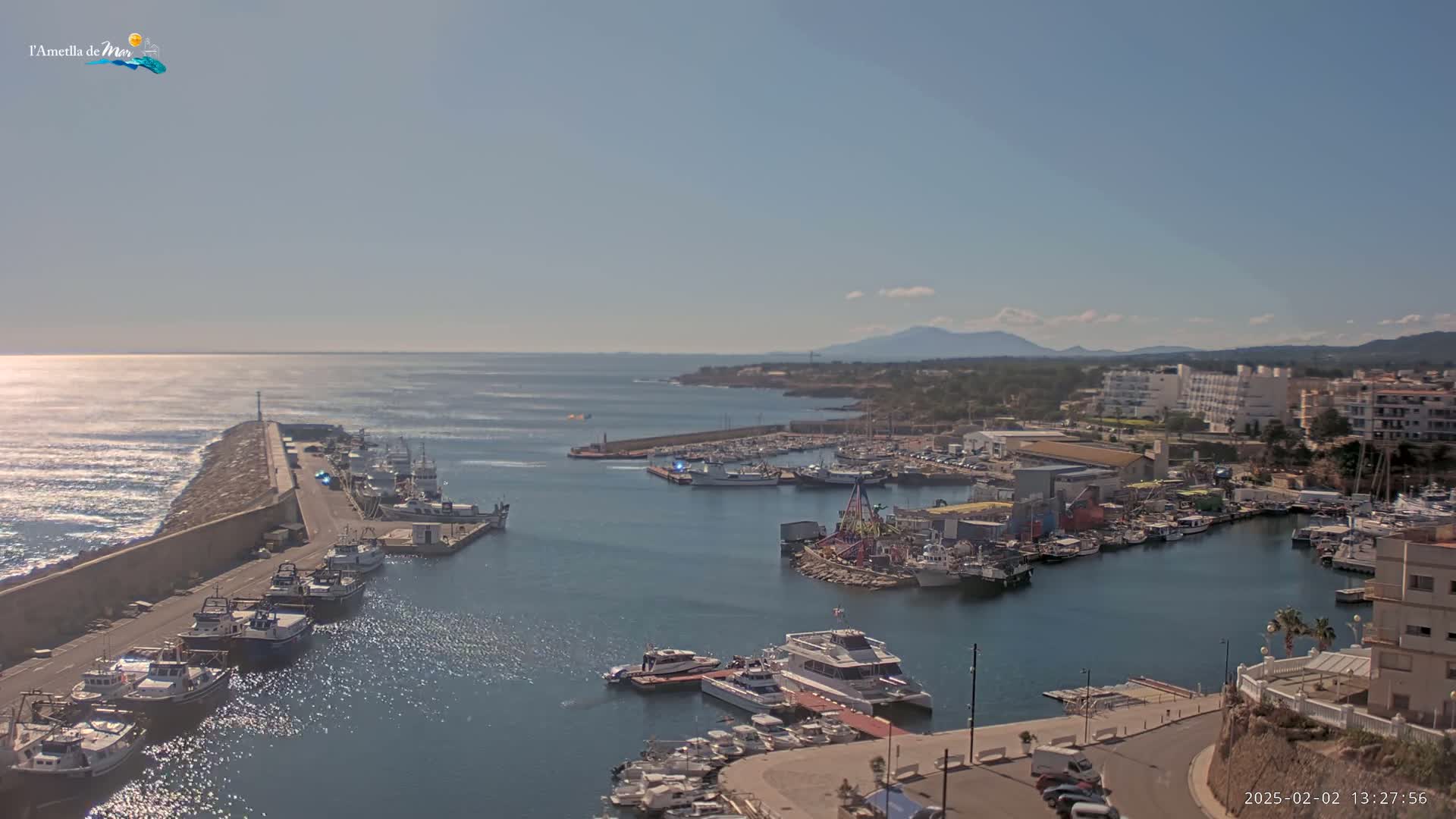 A harbor filled with numerous boats and ships is situated along a coastline under a clear, sunny sky, with mountains visible in the distance.