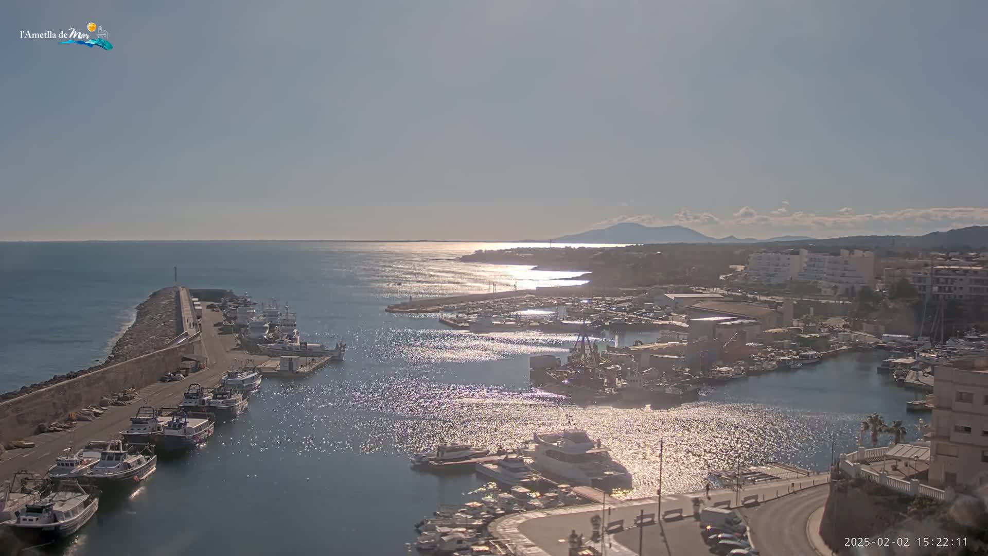 A sunny harbor filled with numerous boats, a stone breakwater, and buildings lining the shoreline, with mountains visible in the distance.