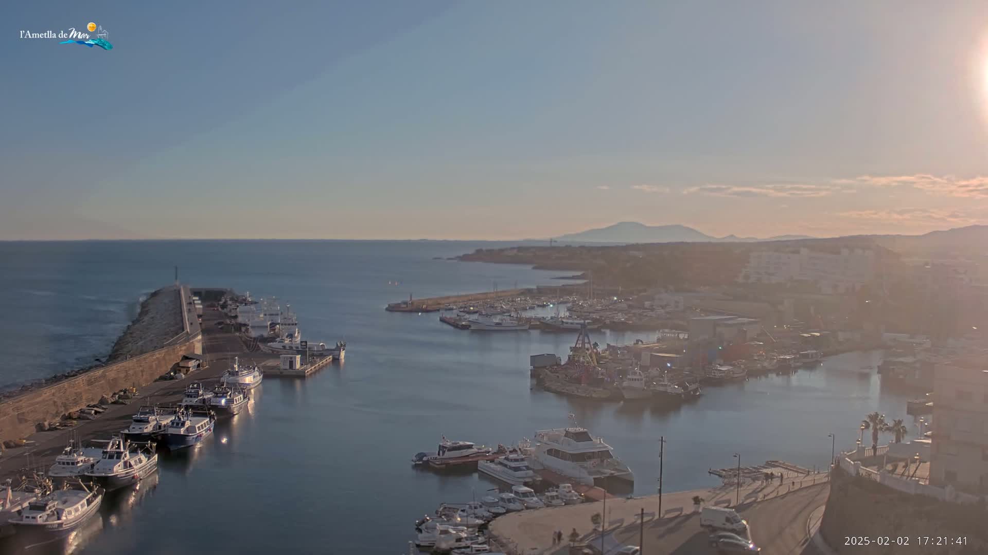 A harbor filled with numerous boats is situated next to a long stone wall, overlooking a calm sea under a clear, slightly hazy sky.