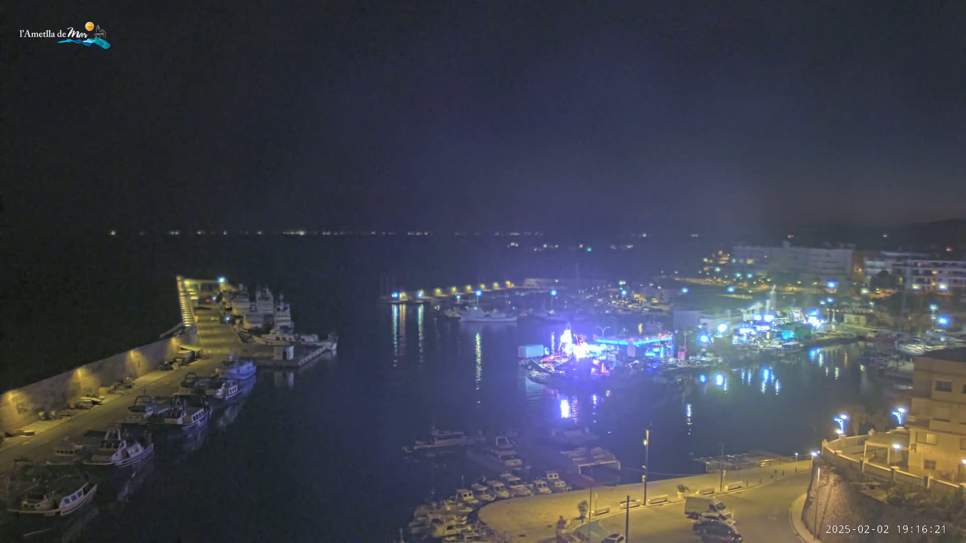 A nighttime view of a harbor filled with boats, illuminated by lights from buildings and a brightly lit area on the waterfront.