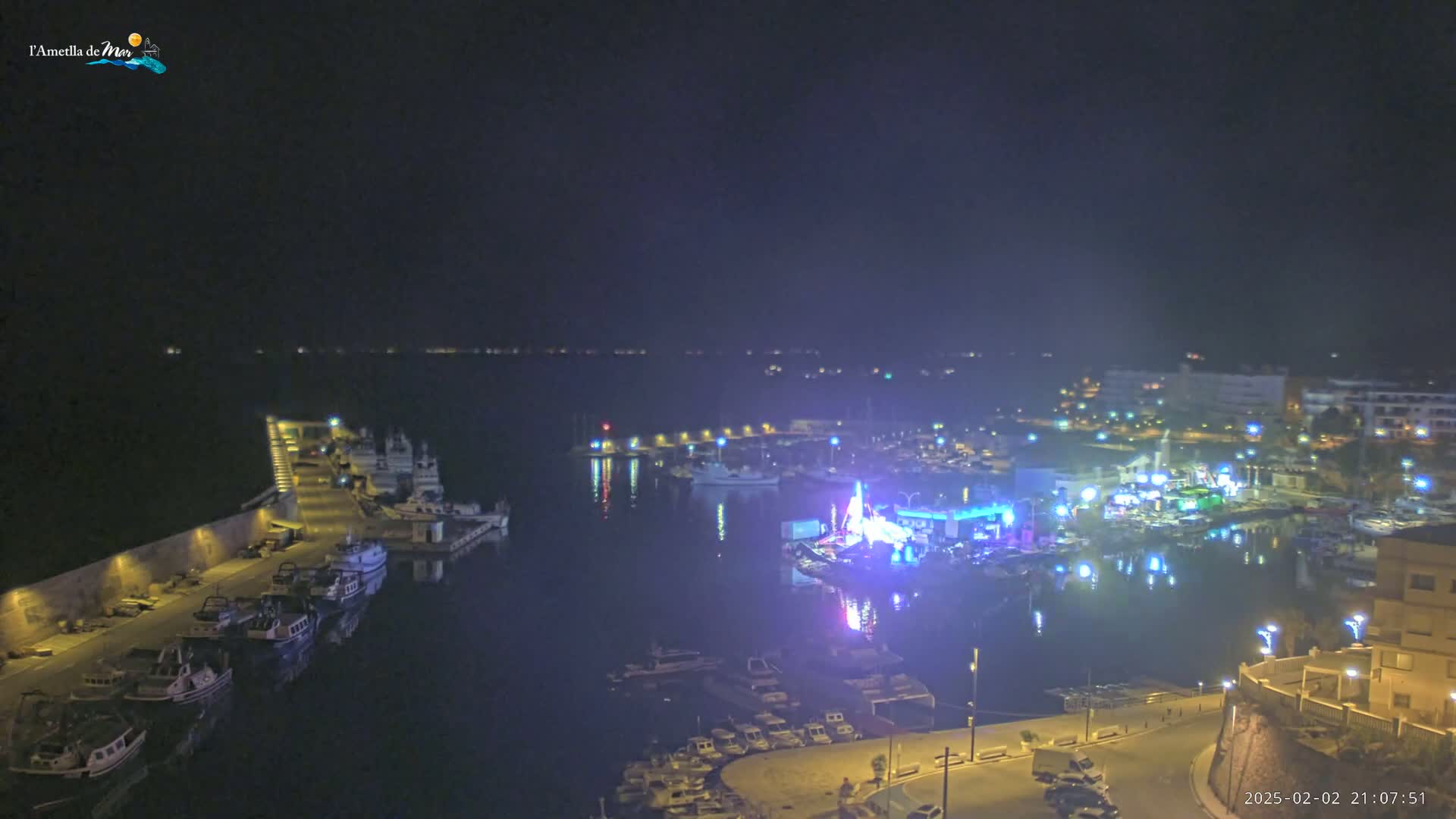 A nighttime view of a harbor filled with boats, illuminated by lights from buildings and a brightly lit structure on the water.