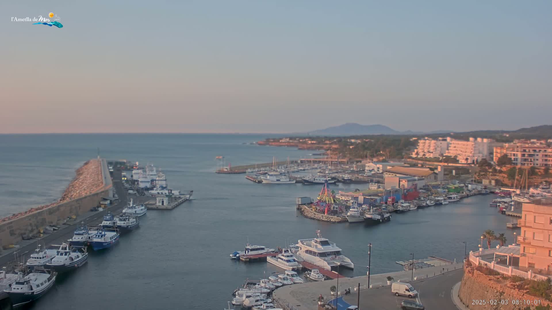 A harbor filled with numerous boats and ships is nestled alongside a coastline under a clear, pale sunset sky.