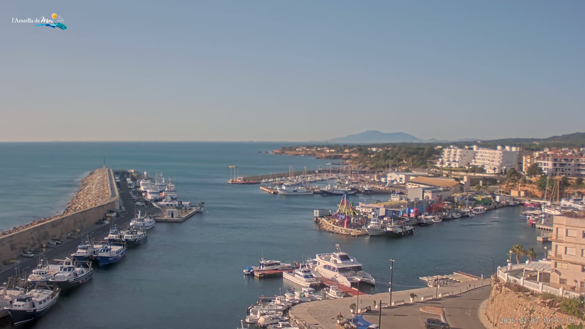A harbor filled with numerous boats and yachts is seen on a sunny day, with calm waters extending to a distant coastline under a clear sky.