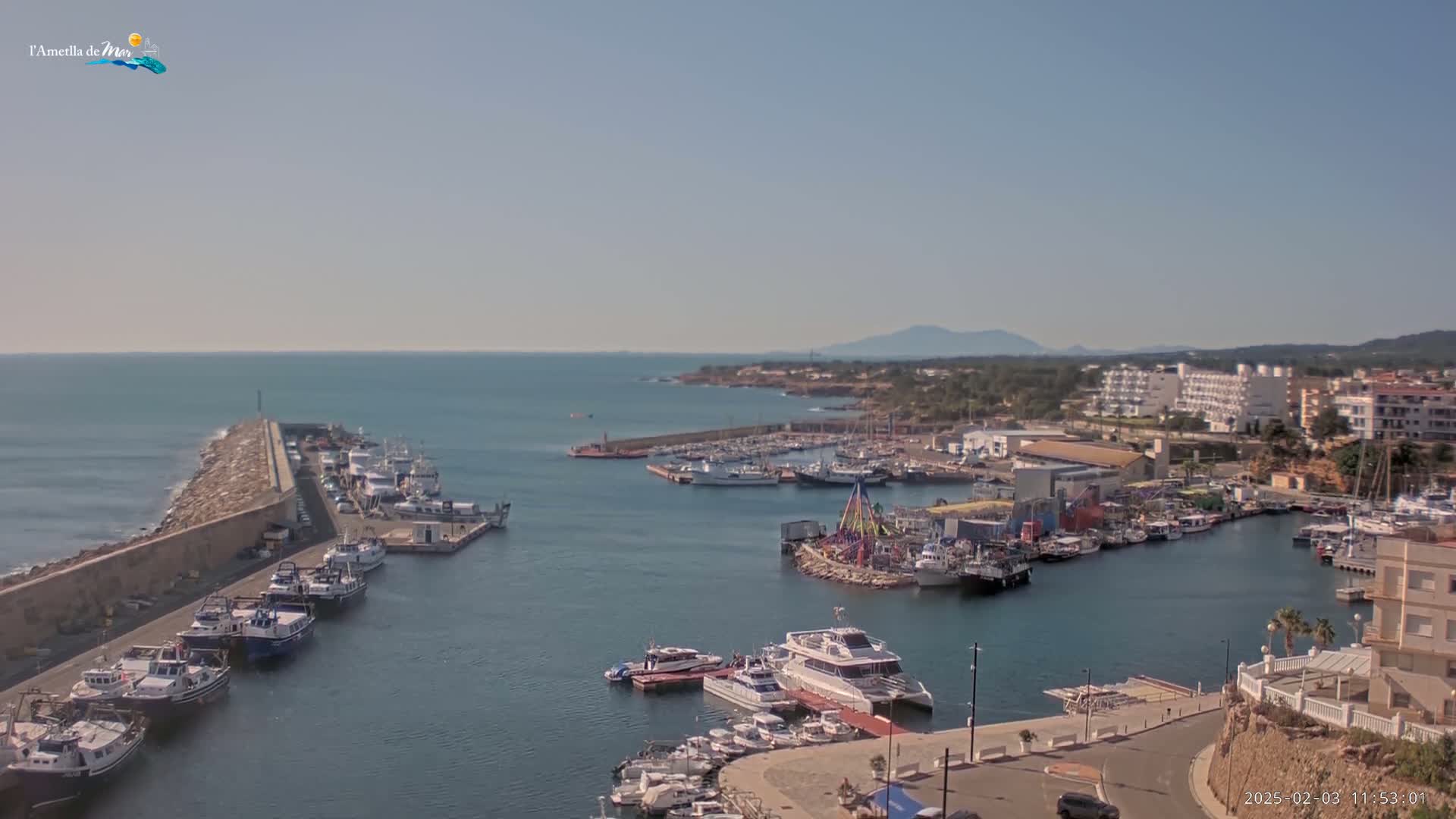 A harbor filled with numerous boats and ships is seen on a sunny day, with calm waters and a clear view of the distant coastline and mountains.