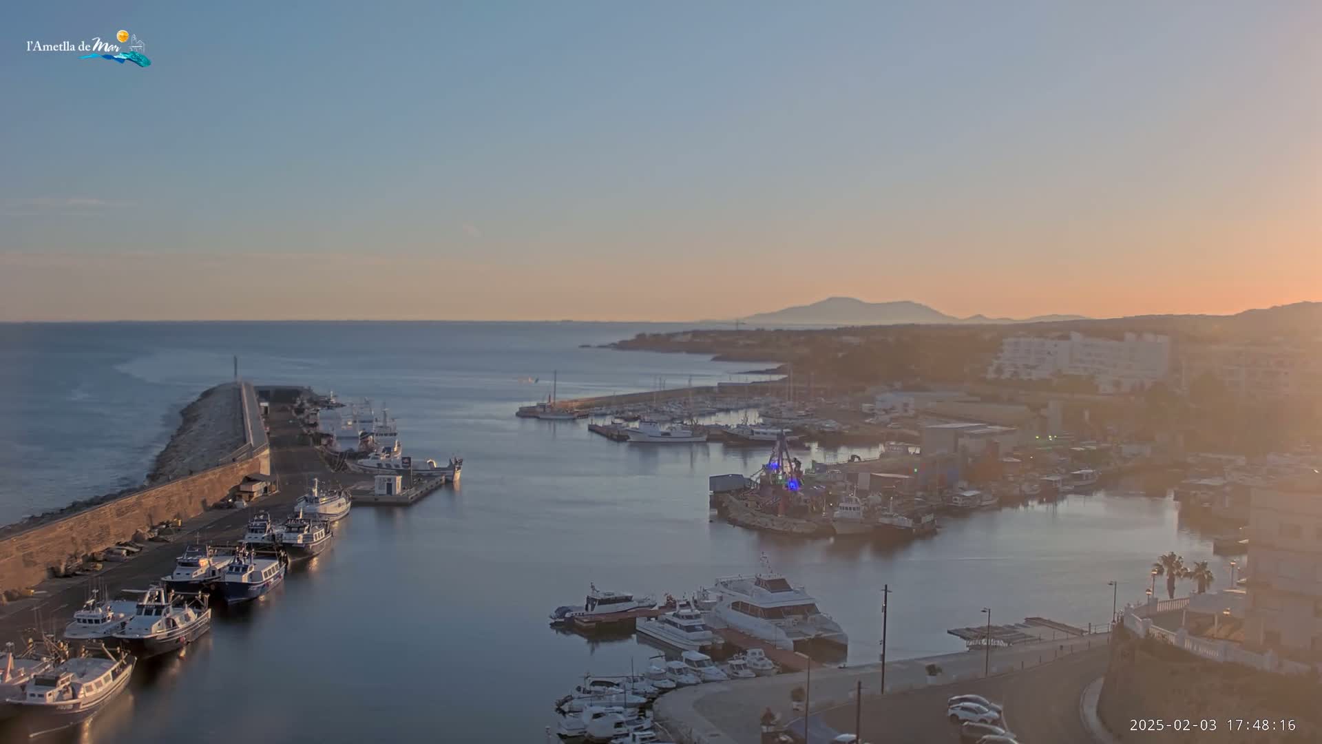 A calm harbor filled with numerous boats is seen at sunset, with a long stone wall extending into the sea.