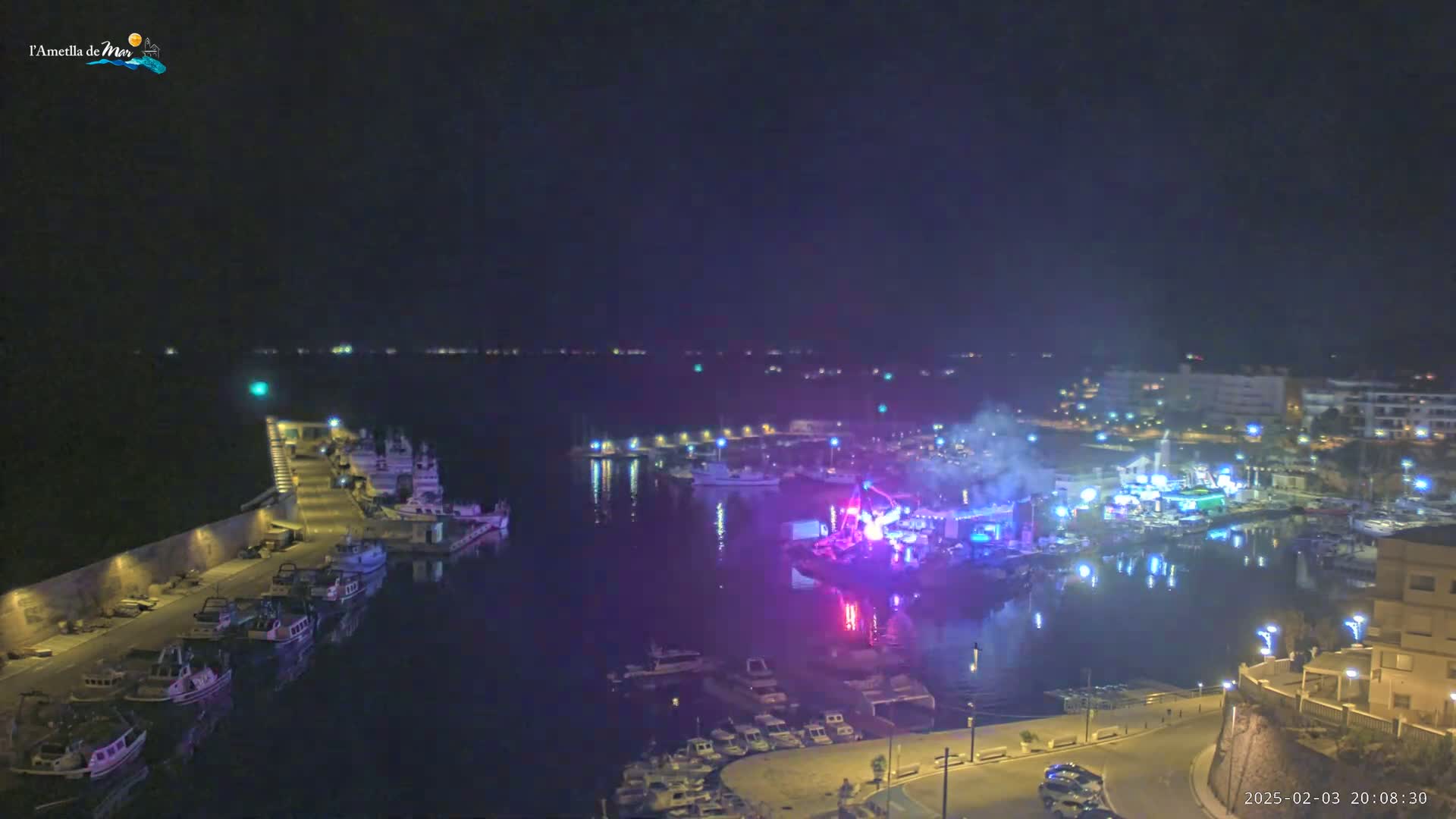 A nighttime aerial view of a harbor filled with boats, with a brightly lit stage and some smoke visible in the water, under a clear, dark sky.