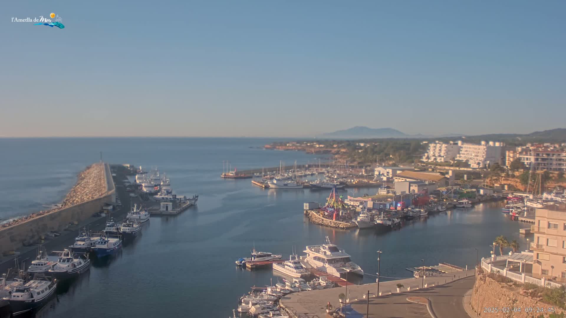 A harbor filled with numerous boats and yachts is nestled along a coastline under a clear, sunny sky.