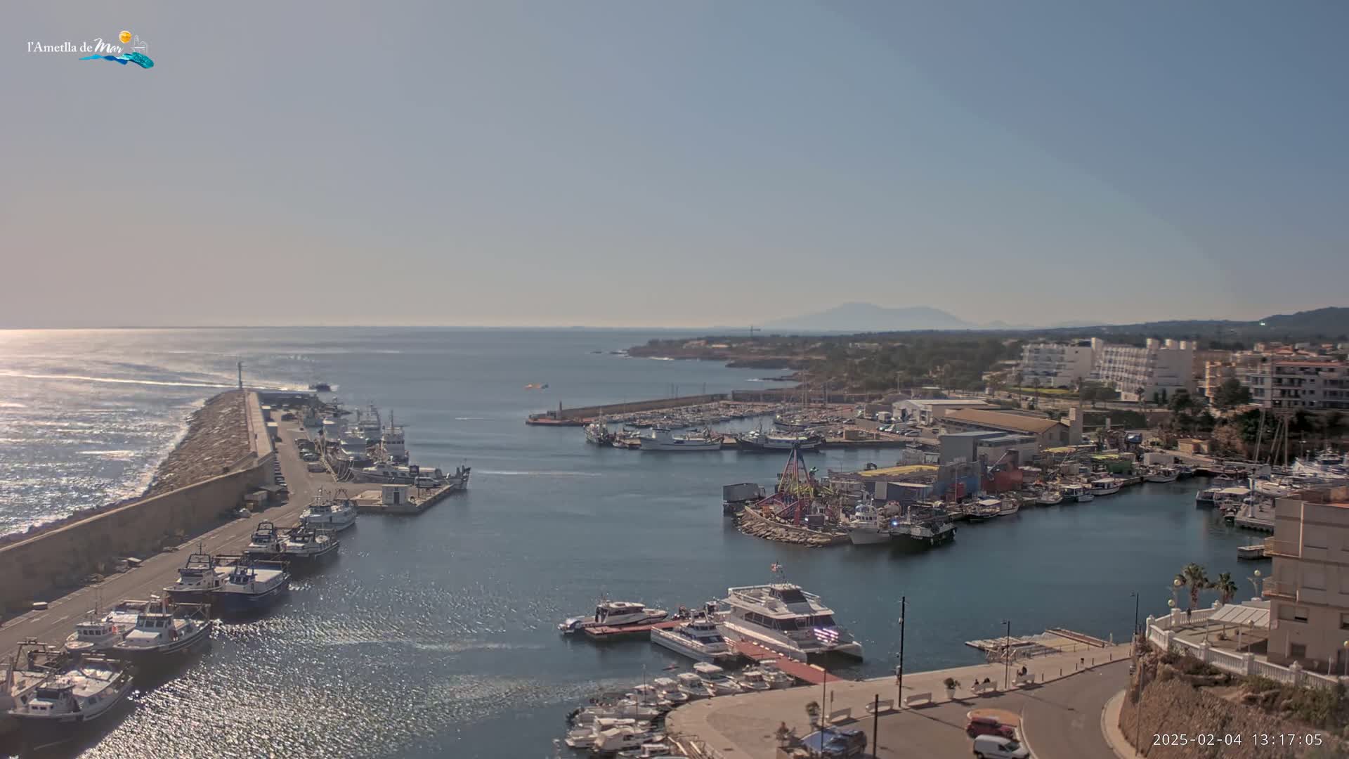 A harbor filled with numerous boats and ships on a sunny day, with calm waters and a coastal town visible in the background.