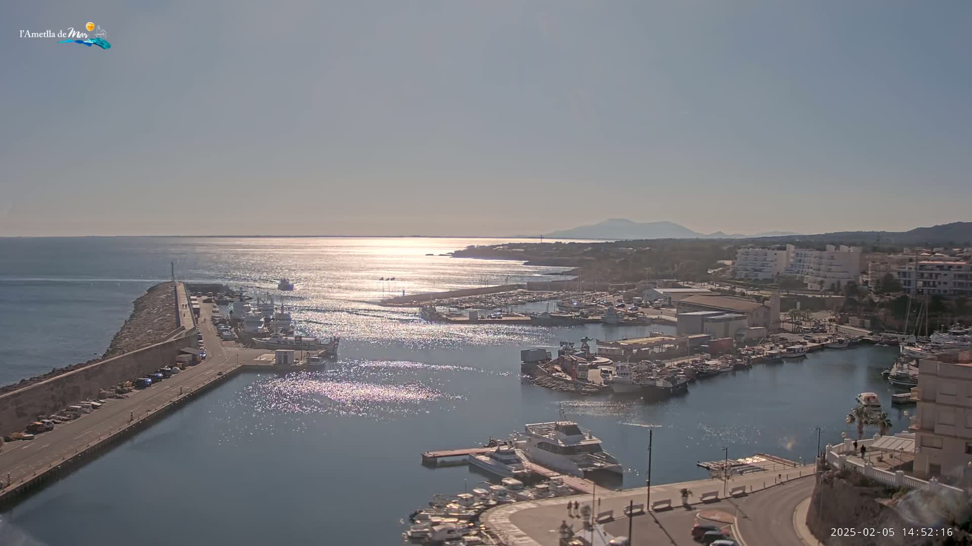 A sunny day reveals a harbor filled with boats, a stone breakwater, and buildings along a coastline under a clear sky.