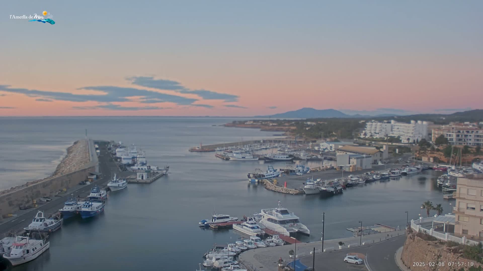 A harbor filled with numerous boats and yachts is shown at dawn under a pale pink and blue sky.