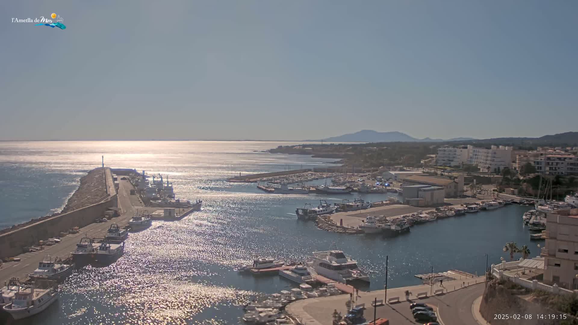 A sunny day shows a harbor filled with many boats of varying sizes, a long stone breakwater, and buildings along the coast, with mountains visible in the distance.