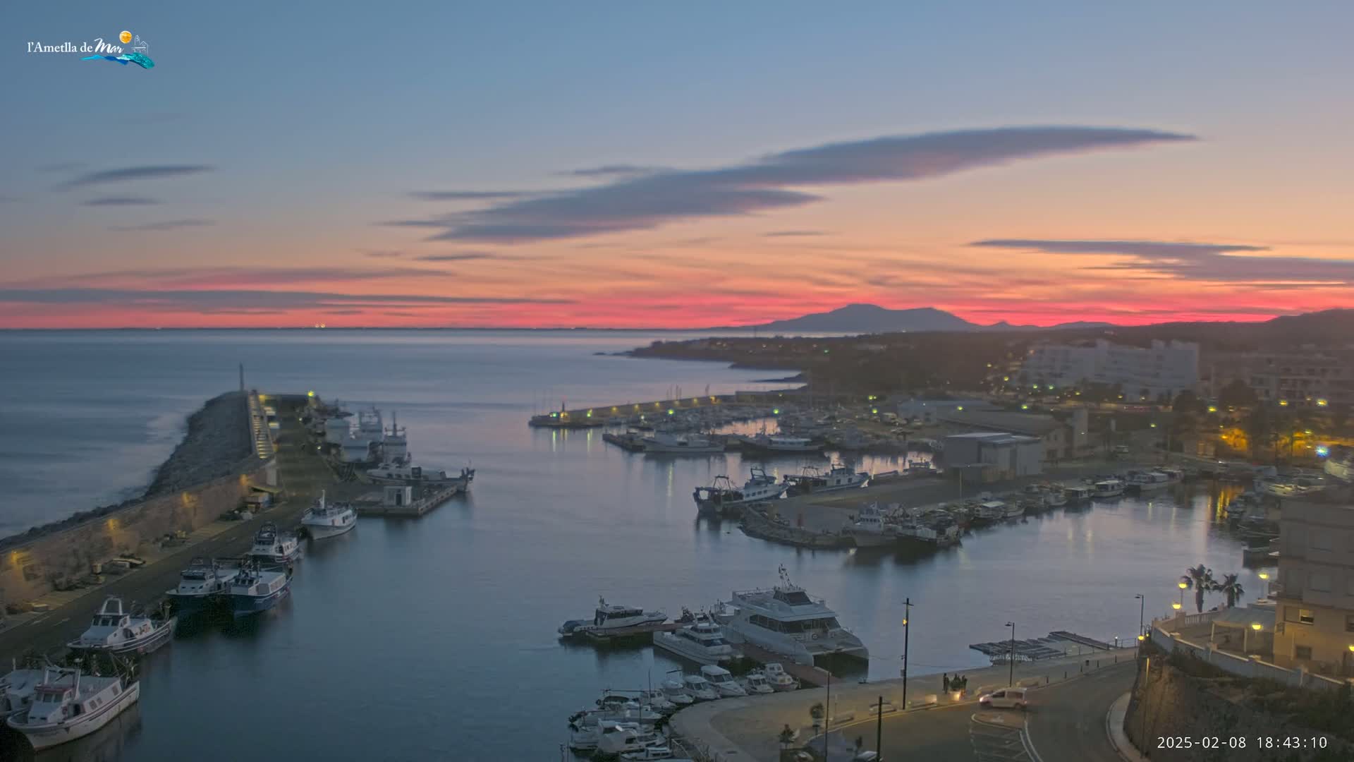 A harbor filled with boats is seen at sunset, with a pink and orange sky above calm water.
