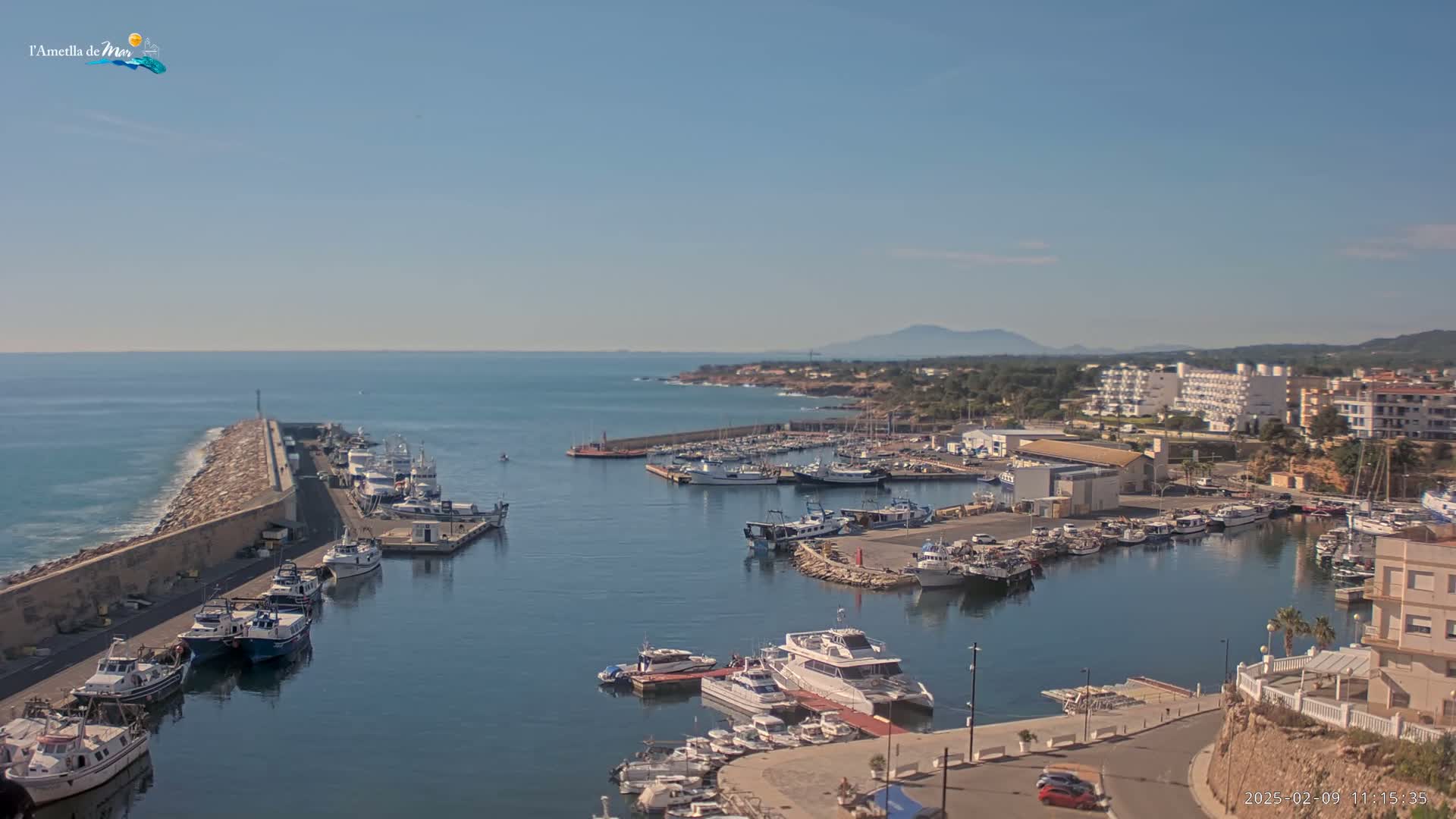 A harbor filled with numerous boats and yachts is situated along a calm, clear ocean under a sunny sky, with a coastal town and distant mountains visible in the background.