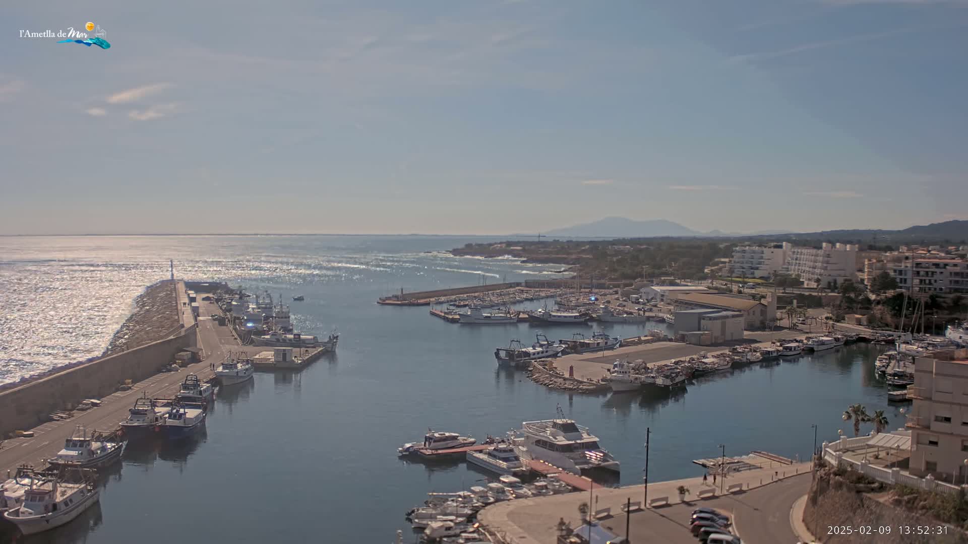 A harbor filled with numerous boats and yachts under a mostly sunny sky, with calm waters and a distant coastline visible.