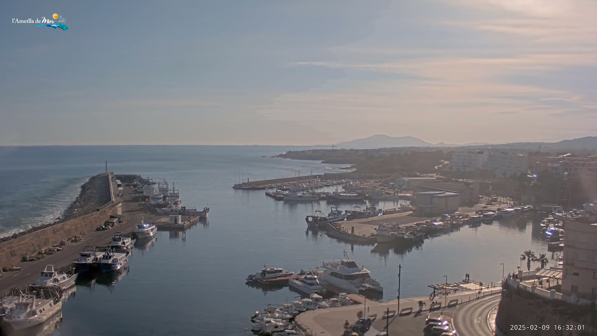 A harbor filled with numerous boats is situated on a calm sea under a partly cloudy sky, with distant mountains visible on the horizon.