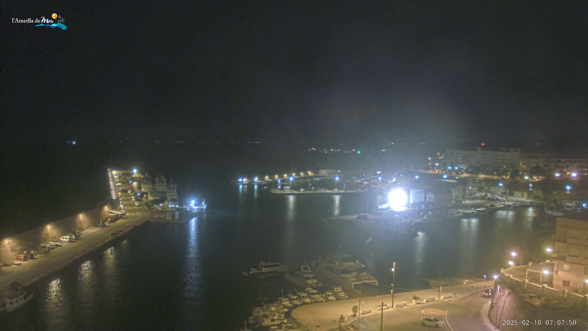 A nighttime, aerial view of a harbor filled with boats, illuminated by artificial lights under a hazy sky.